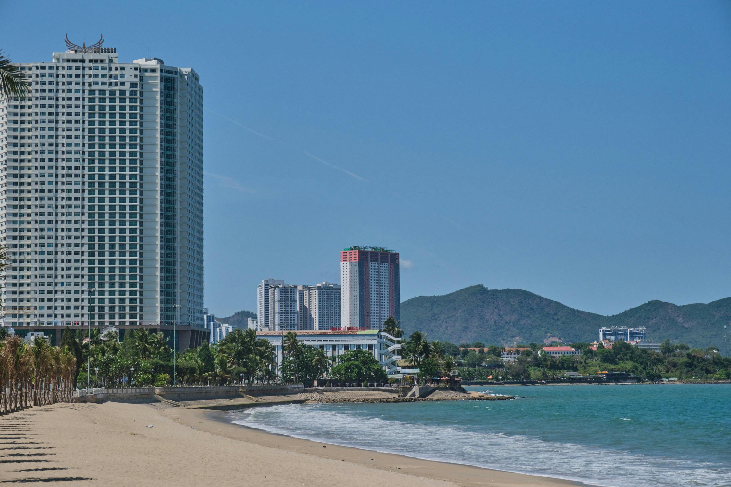 A bustling Nha Trang Beach during daytime with sunbathers, umbrellas, and the turquoise sea, backed by the city skyline and mountains in the distance.