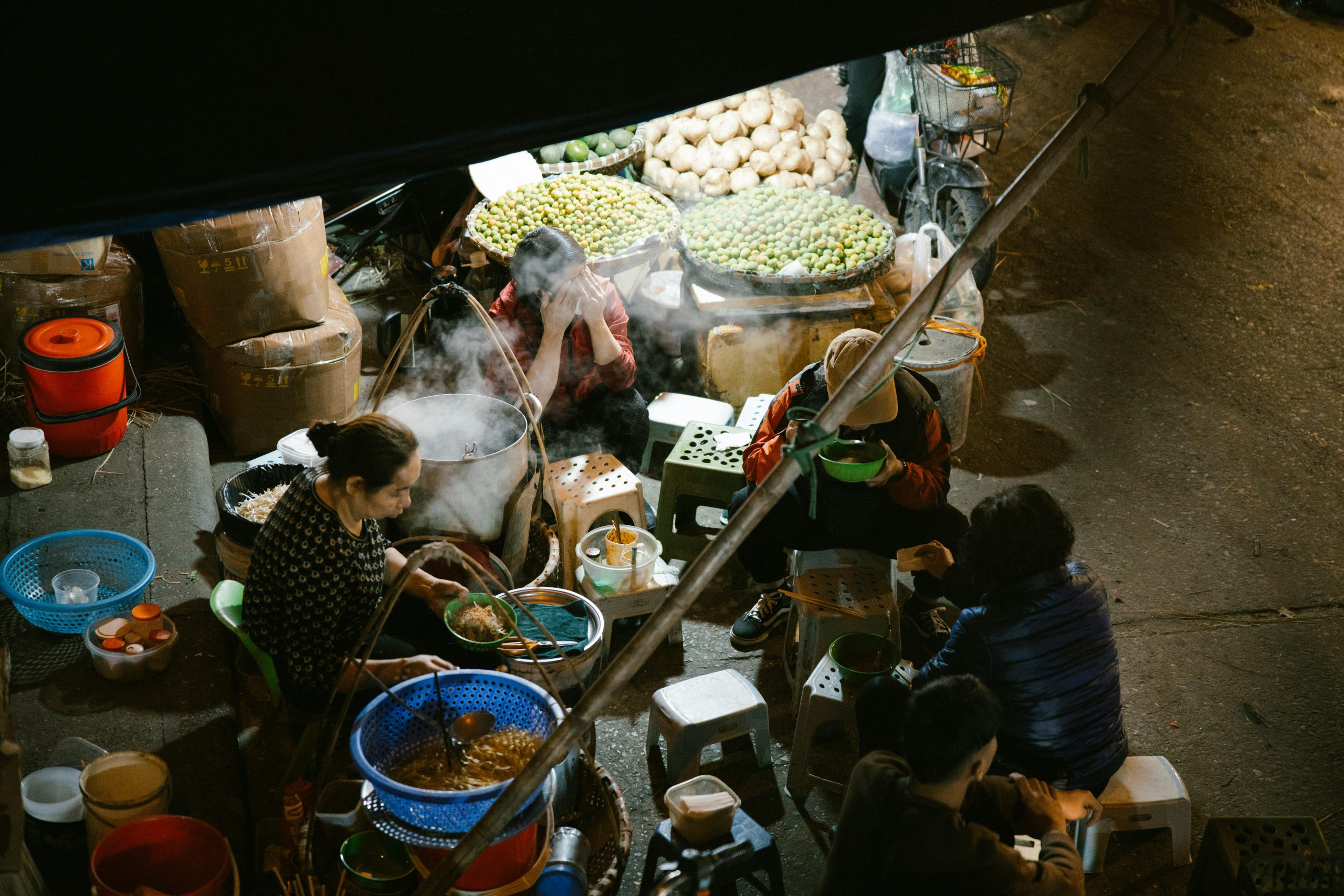 A bustling street food stall in Hanoi's Ba Dinh District during evening, selling traditional pho, with steam rising from bowls and locals enjoying their meal