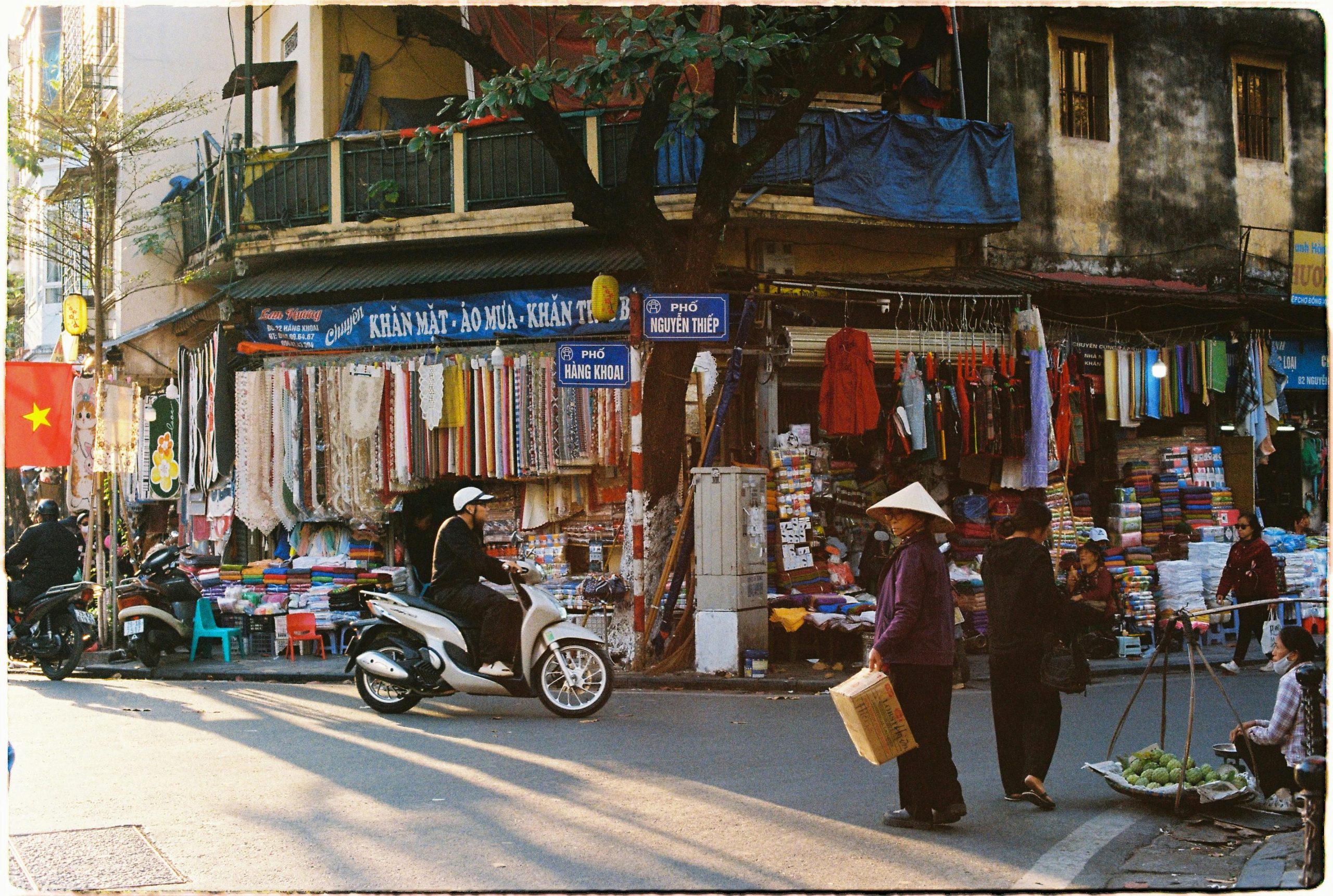 A bustling street scene in Hanoi's Old Quarter with people on motorbikes, street vendors, and traditional shop houses under a warm, hazy sky
