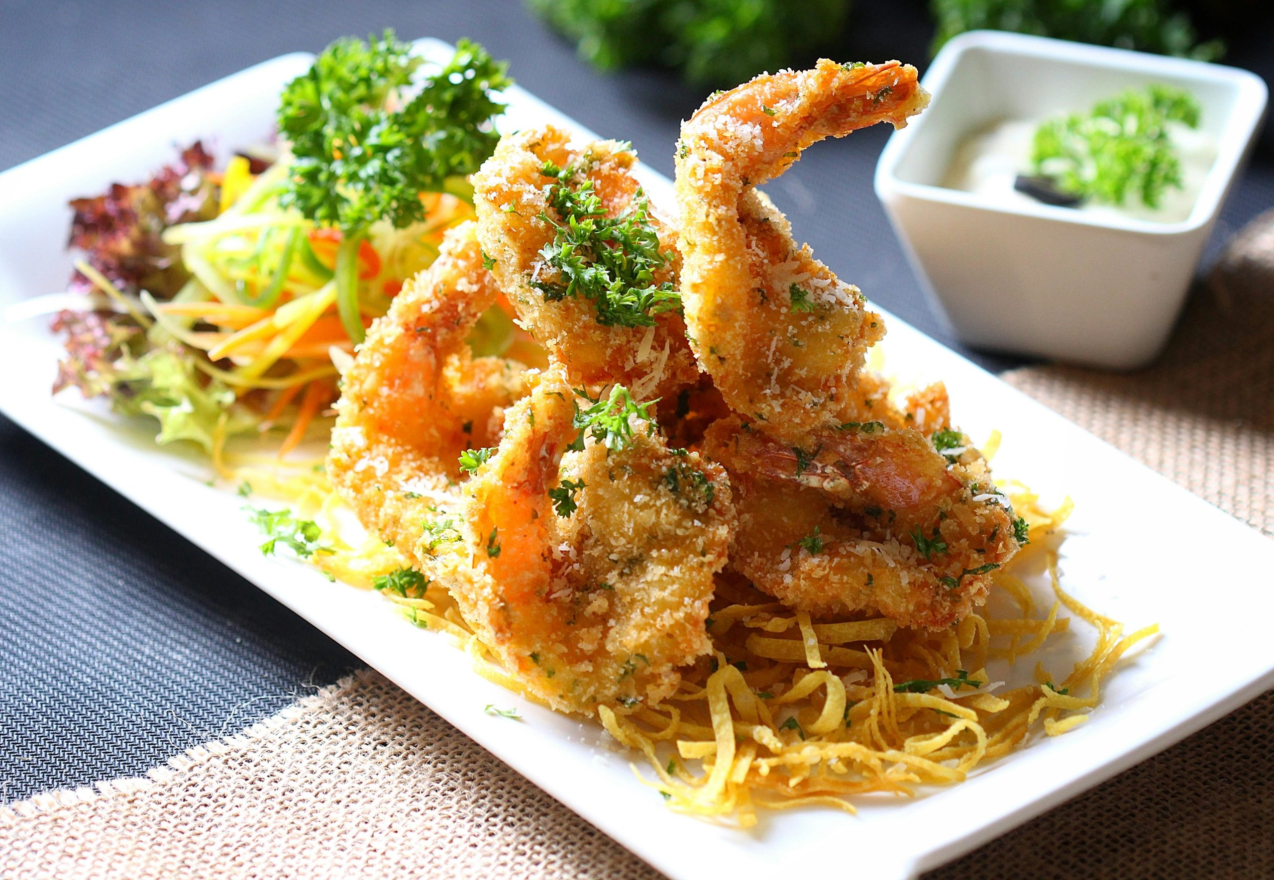 A close-up shot of crispy and golden 'Banh Tom' (shrimp cakes) served with fresh green herbs and a sweet and sour dipping sauce, on a rustic plate in a street food setting near West Lake.