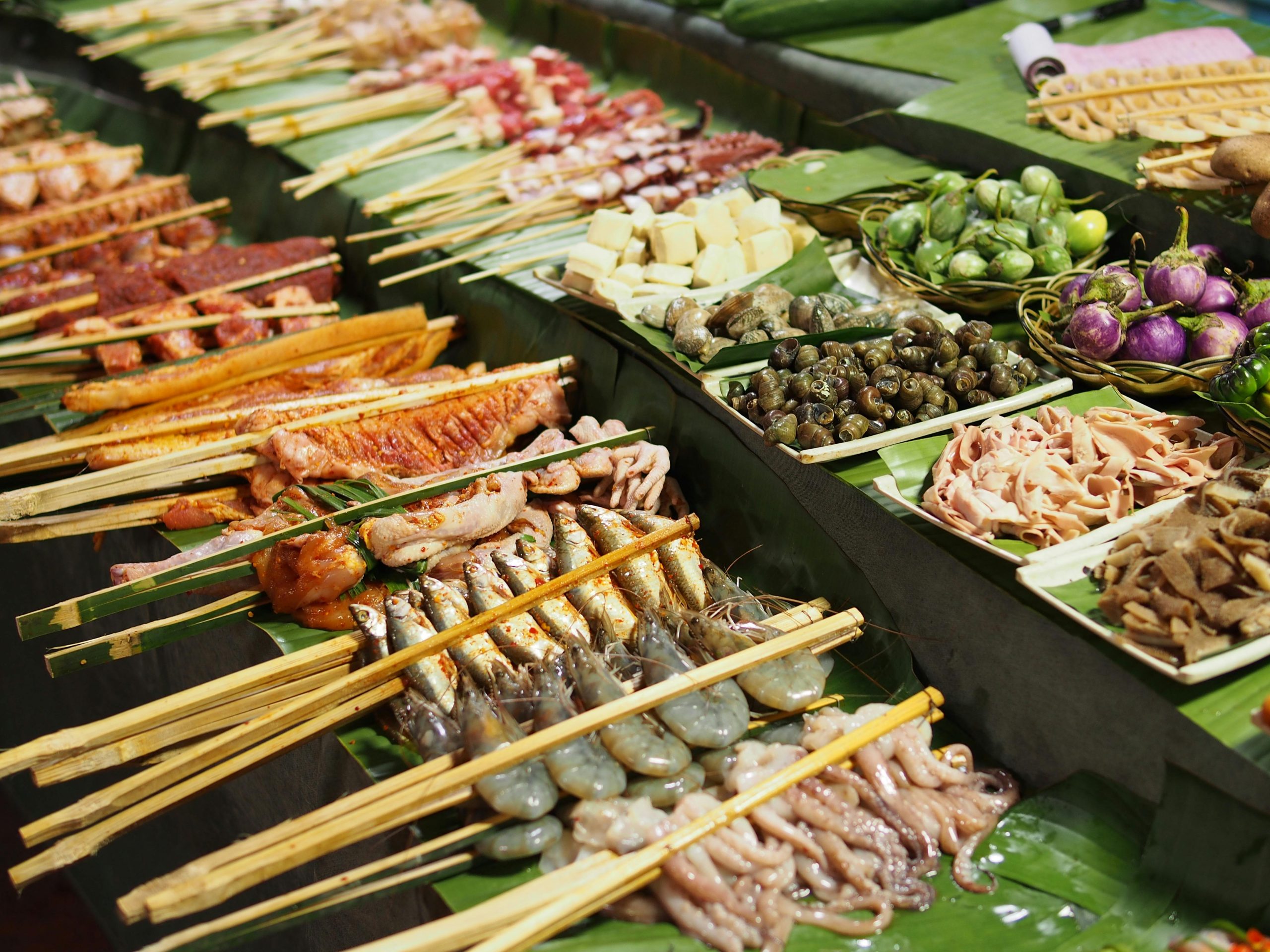 A colorful spread of freshly grilled Vietnamese seafood laid out on a banana leaf on a rustic wooden table by the beach. The dishes include grilled whole fish, large prawns, squid, and various fresh herbs and dipping sauces. The ocean is blurred in the background.