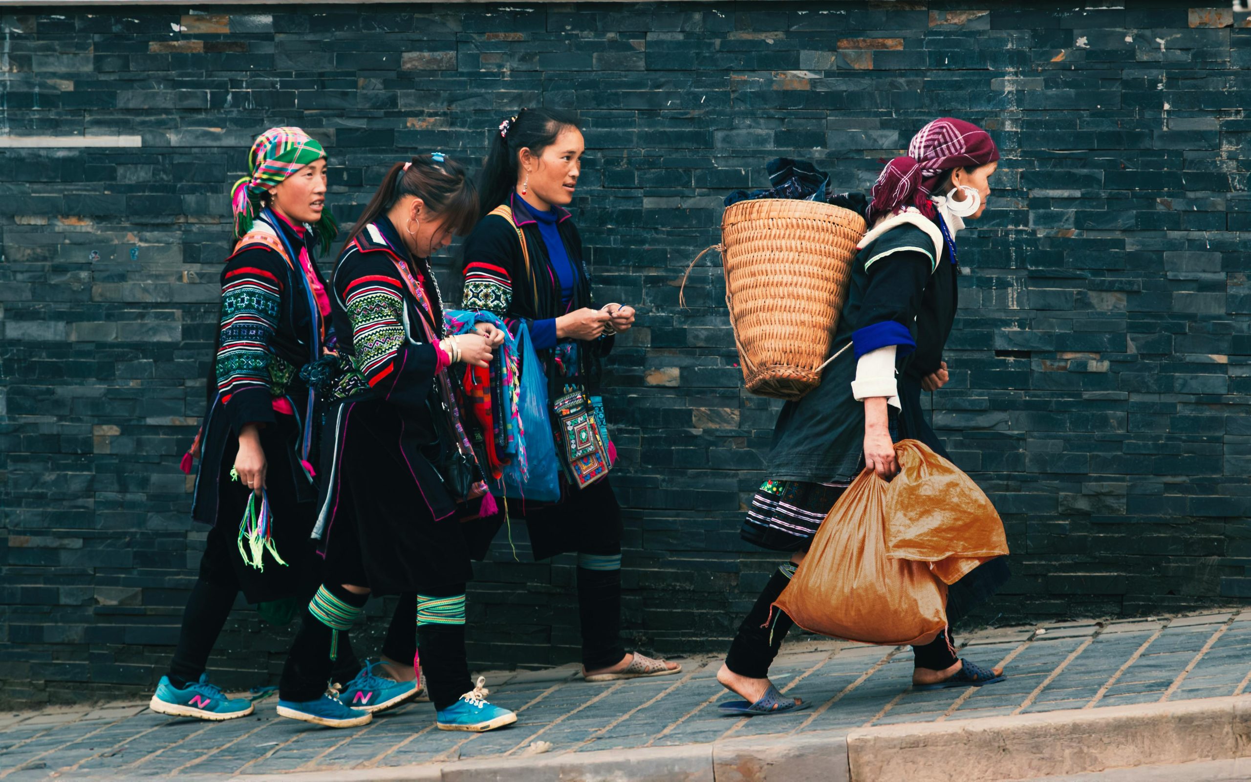 A group of colorful ethnic minority women from the H'mong or Red Dao tribe in traditional indigo and embroidered clothing, smiling and interacting with tourists on a muddy village path in Muong Hoa Valley, with terraced fields in the background
