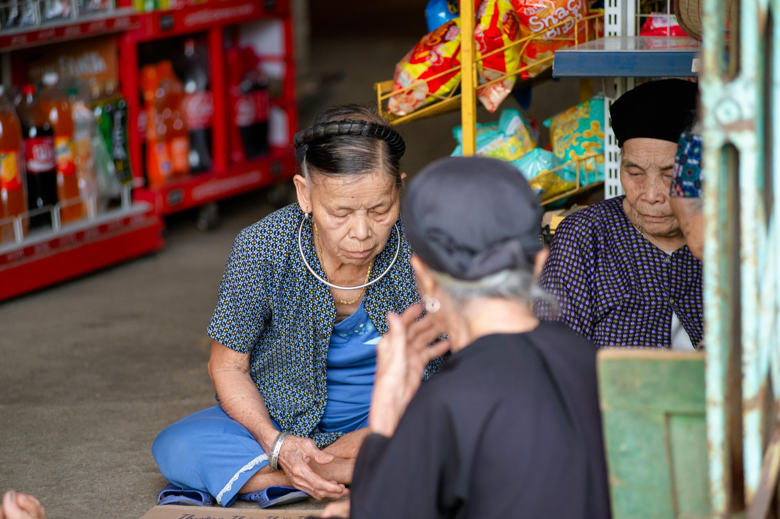 A group of ethnic minority women in traditional colorful attire, smiling and chatting at a bustling local market in Ha Giang, with fresh produce and handicrafts displayed around them