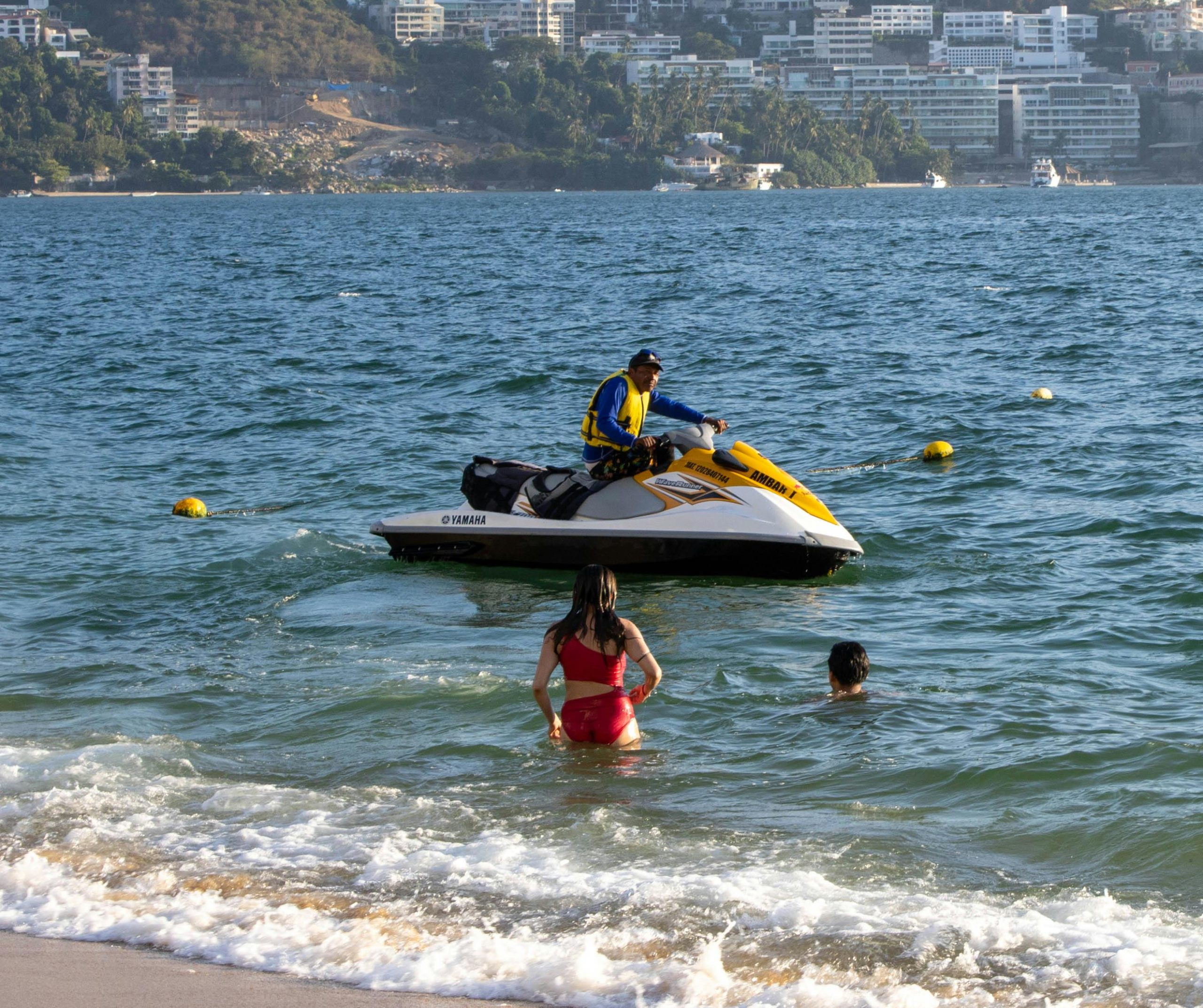 A group of tourists and locals enjoying various water sports at My Khe Beach, including people on jet skis leaving white wakes, a person parasailing high above the ocean, and others swimming and playing in the gentle waves. The background shows the vibrant Da Nang cityscape.