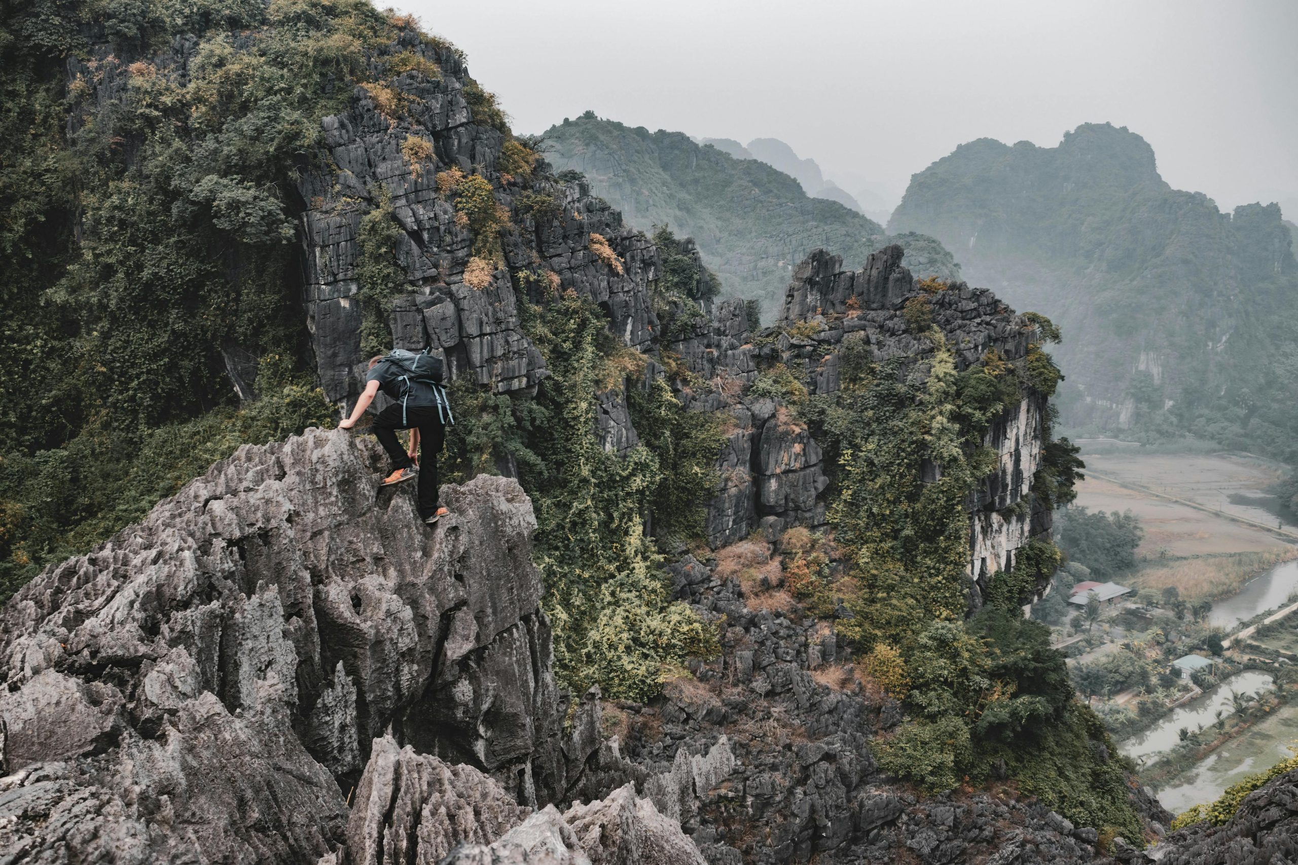 A hiker stands triumphantly at the summit of Ngu Lam Peak in Cat Ba National Park, overlooking a vast expanse of lush green jungle and distant limestone karsts of Lan Ha Bay under a hazy sky