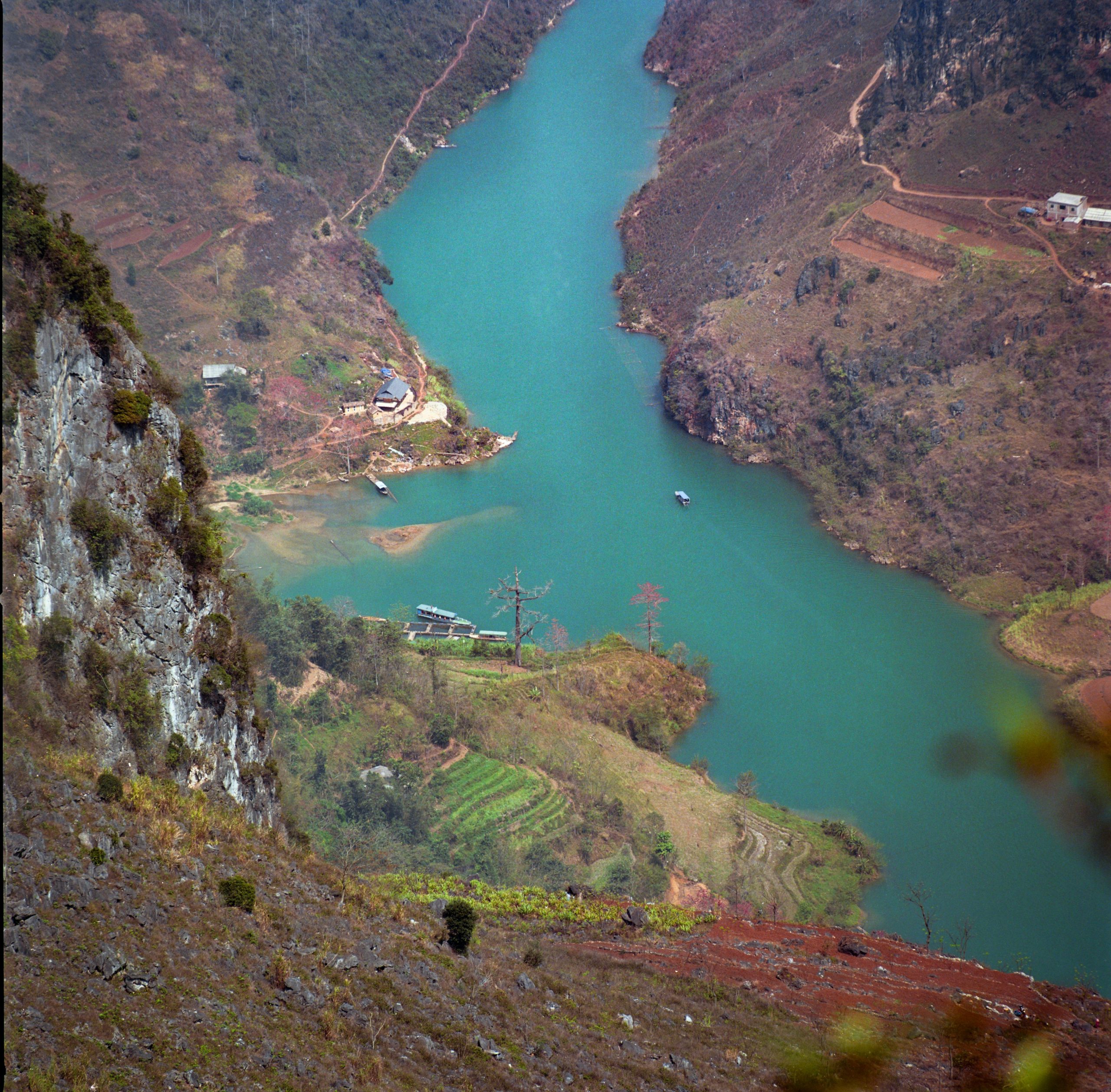 A long, winding road of the Ma Pi Leng Pass carved into a steep cliffside, with a deep green river (Nho Que River) snaking through a dramatic canyon far below, under a clear blue sky