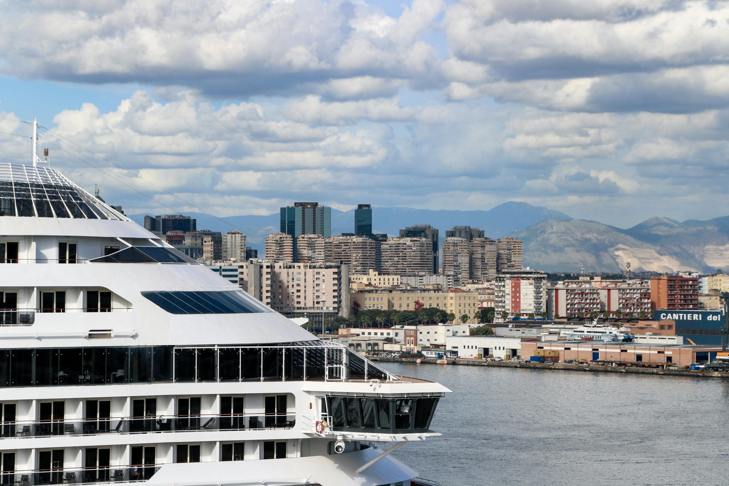 A luxury cruise ship departing from Tuan Chau International Port with the modern port buildings and surrounding hills visible in the background under a cloudy sky