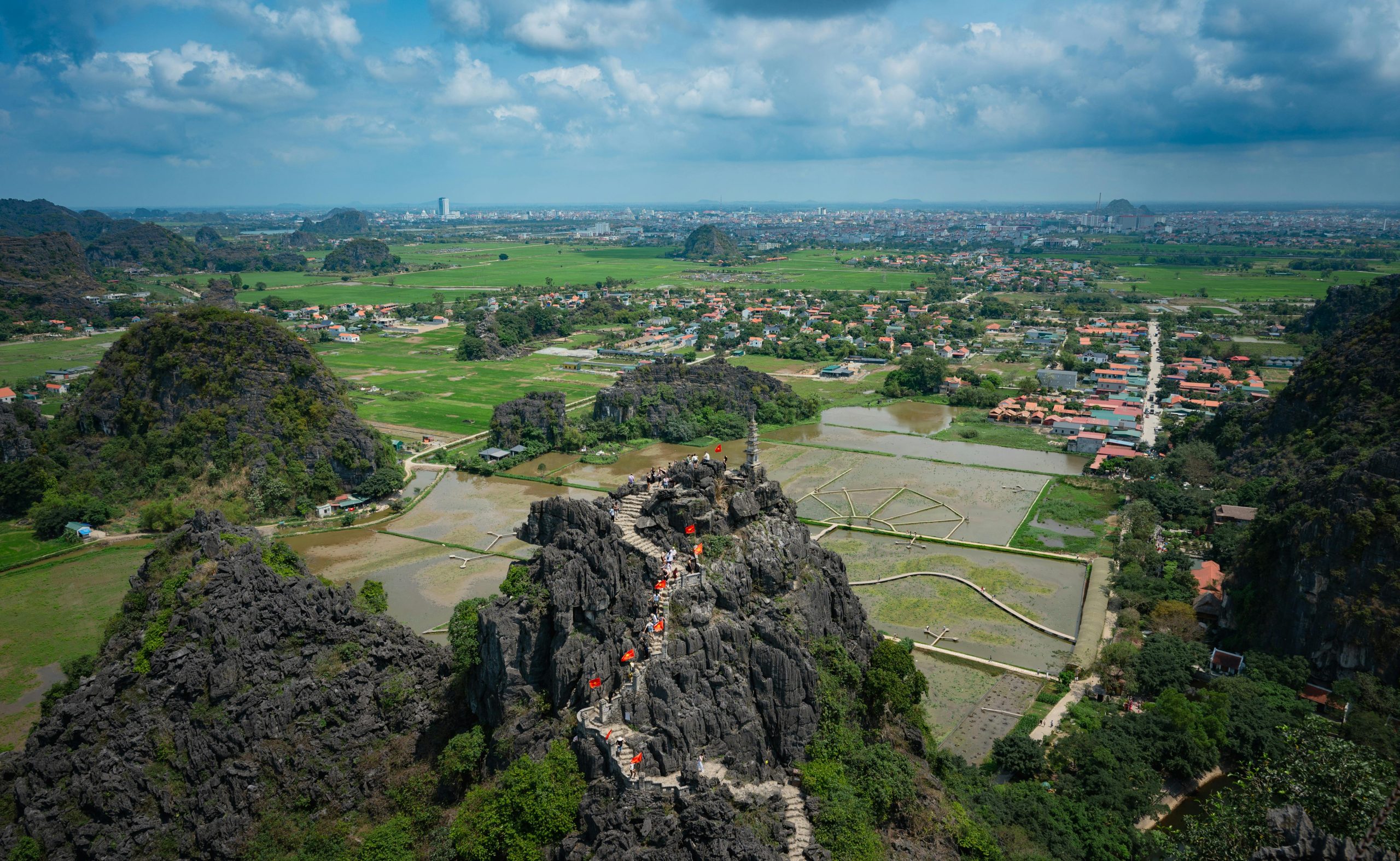 A panoramic aerial view from the Mua Cave viewpoint in Tam Coc, showing vast green rice paddies, the winding Ngo Dong River, and countless limestone karsts stretching to the horizon under a soft morning light.
