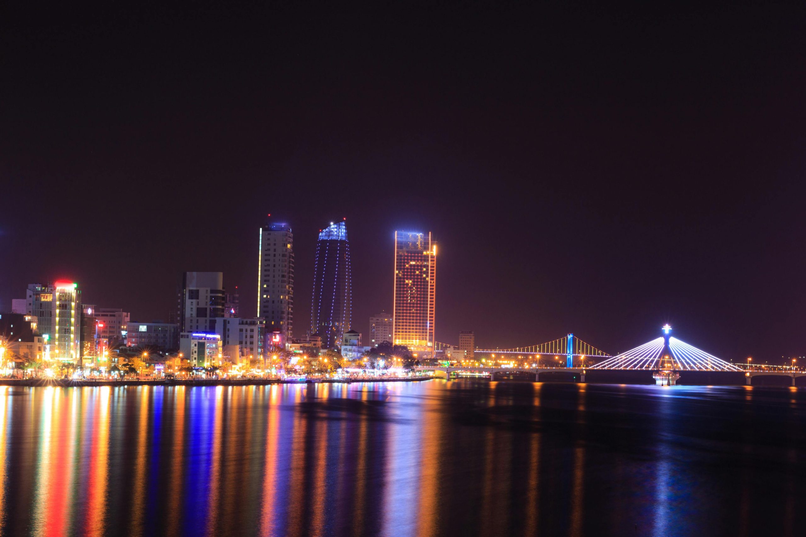 A panoramic view of Da Nang city skyline at night with the lit-up bridges over the Han River, including the Dragon Bridge and Tran Thi Ly Bridge, reflecting on the water, against a backdrop of city lights and mountains