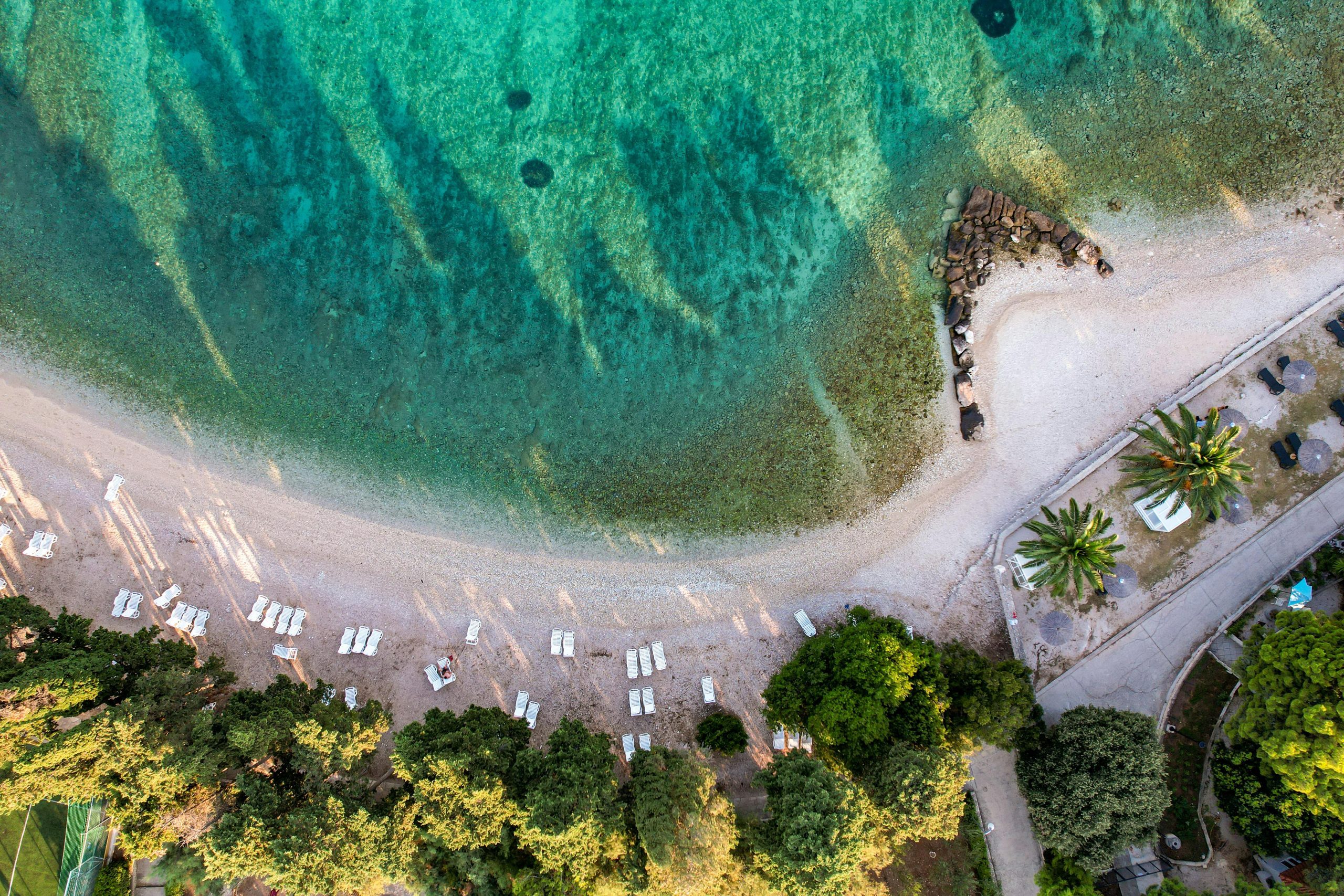 A panoramic view of the wide, golden crescent of My Khe Beach with clear turquoise water, gentle waves, and lush green palm trees in the foreground. Several colorful parasols and sun loungers are visible, with the distant Da Nang skyline under a bright blue sky.