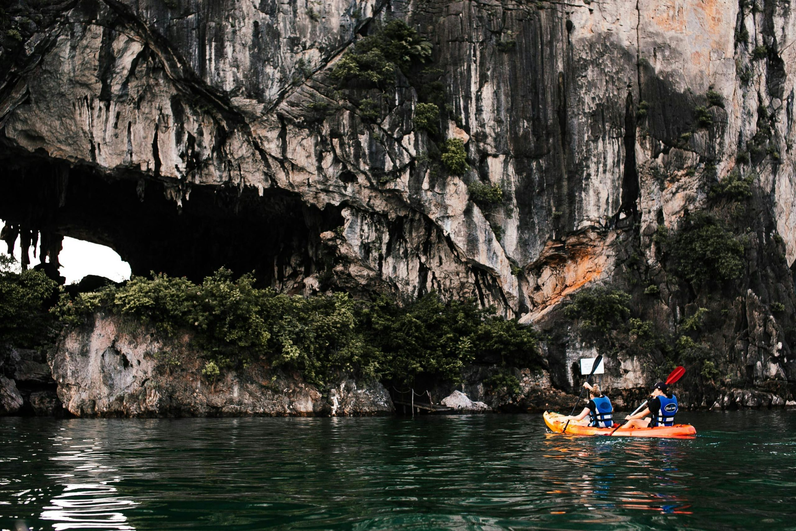 A person in a red kayak paddles through a narrow, dark cave entrance, revealing a brightly lit, secluded emerald lagoon beyond, surrounded by towering, lush green limestone cliffs in Ha Long Bay