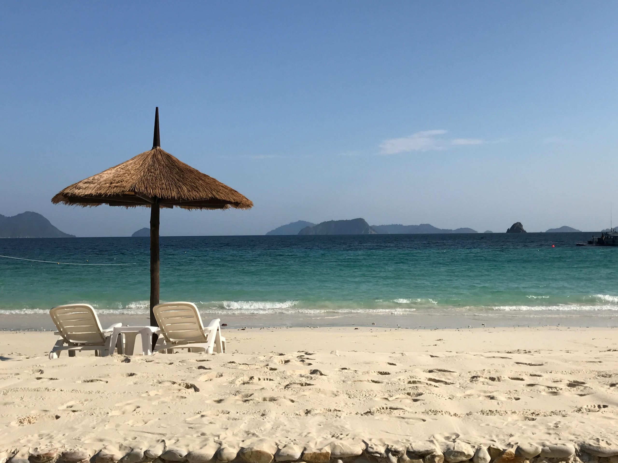 A picturesque view of An Bang Beach with empty bamboo beach loungers and vibrant blue umbrellas set up on soft golden sand, facing the calm turquoise ocean under a clear sunny sky. In the background, lush green palm trees line the shore.