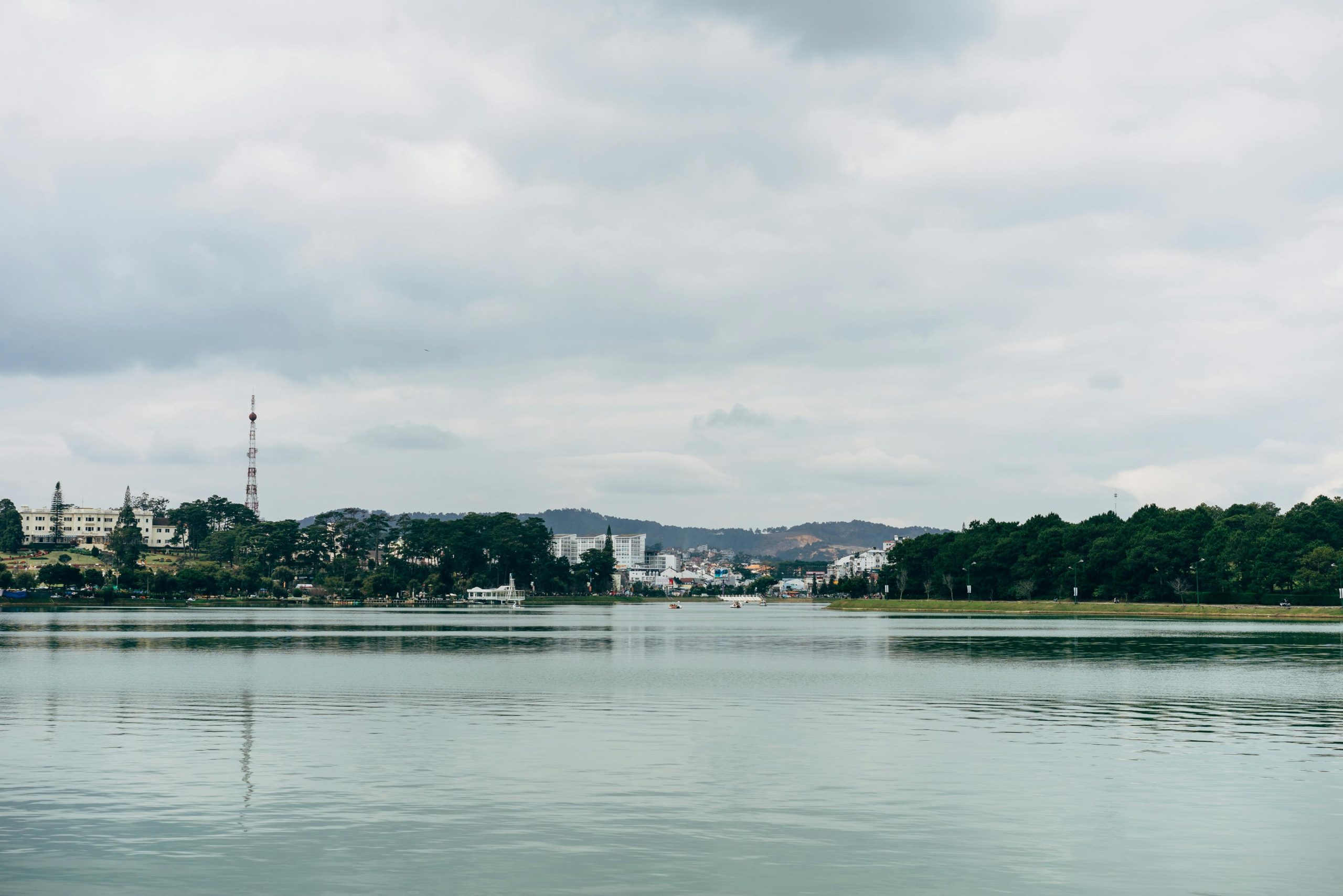 A scenic view of Xuan Huong Lake in Da Lat, Vietnam, with paddle boats on the calm water, surrounded by vibrant flower gardens and pine trees, with colonial-era buildings visible in the distance under a partly cloudy sky.