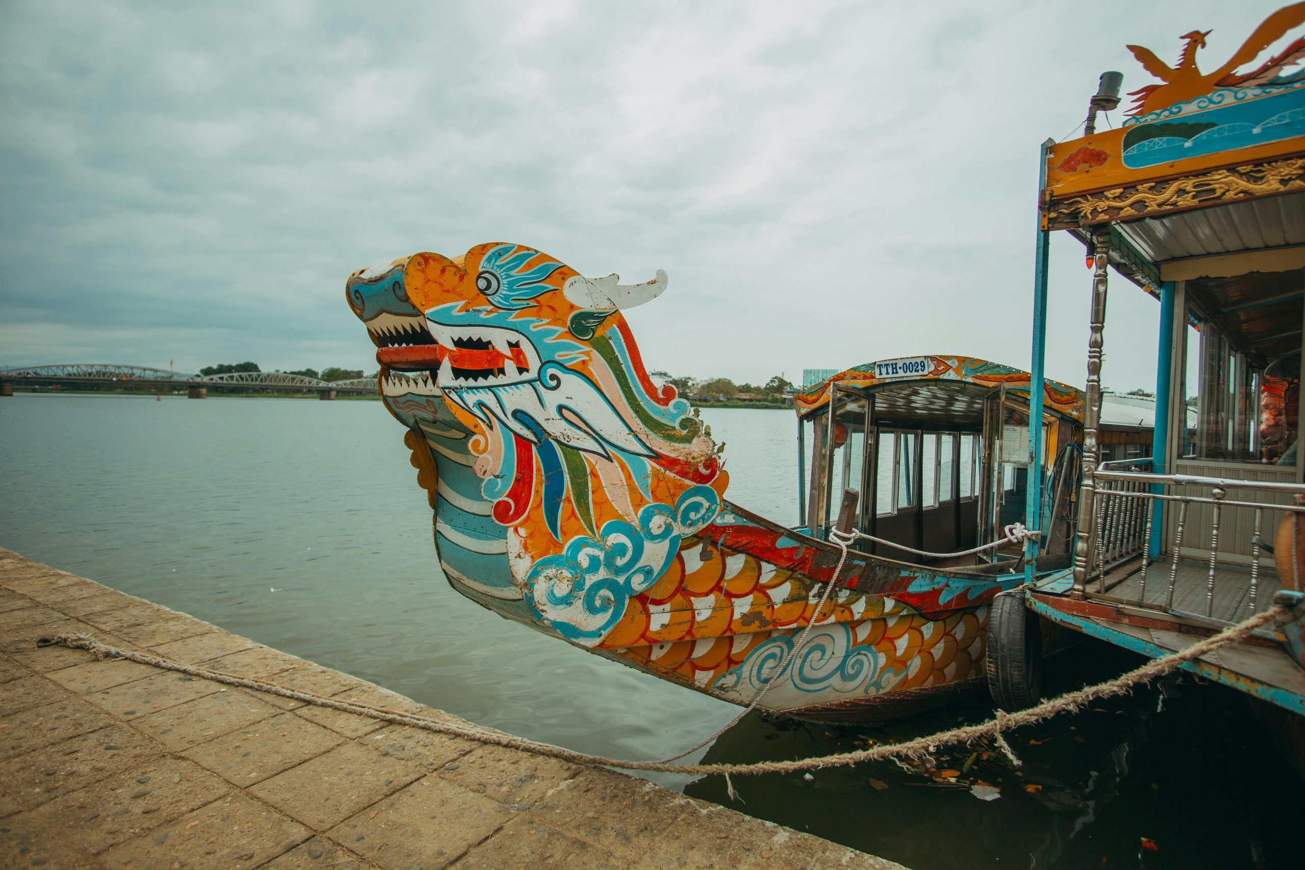 A traditional dragon boat with a curved roof and colorful decorations, cruising slowly on the calm, reflective waters of the Perfume River in Hue. The riverbanks are lined with lush green trees, and the sky is a soft blue with a few scattered clouds.