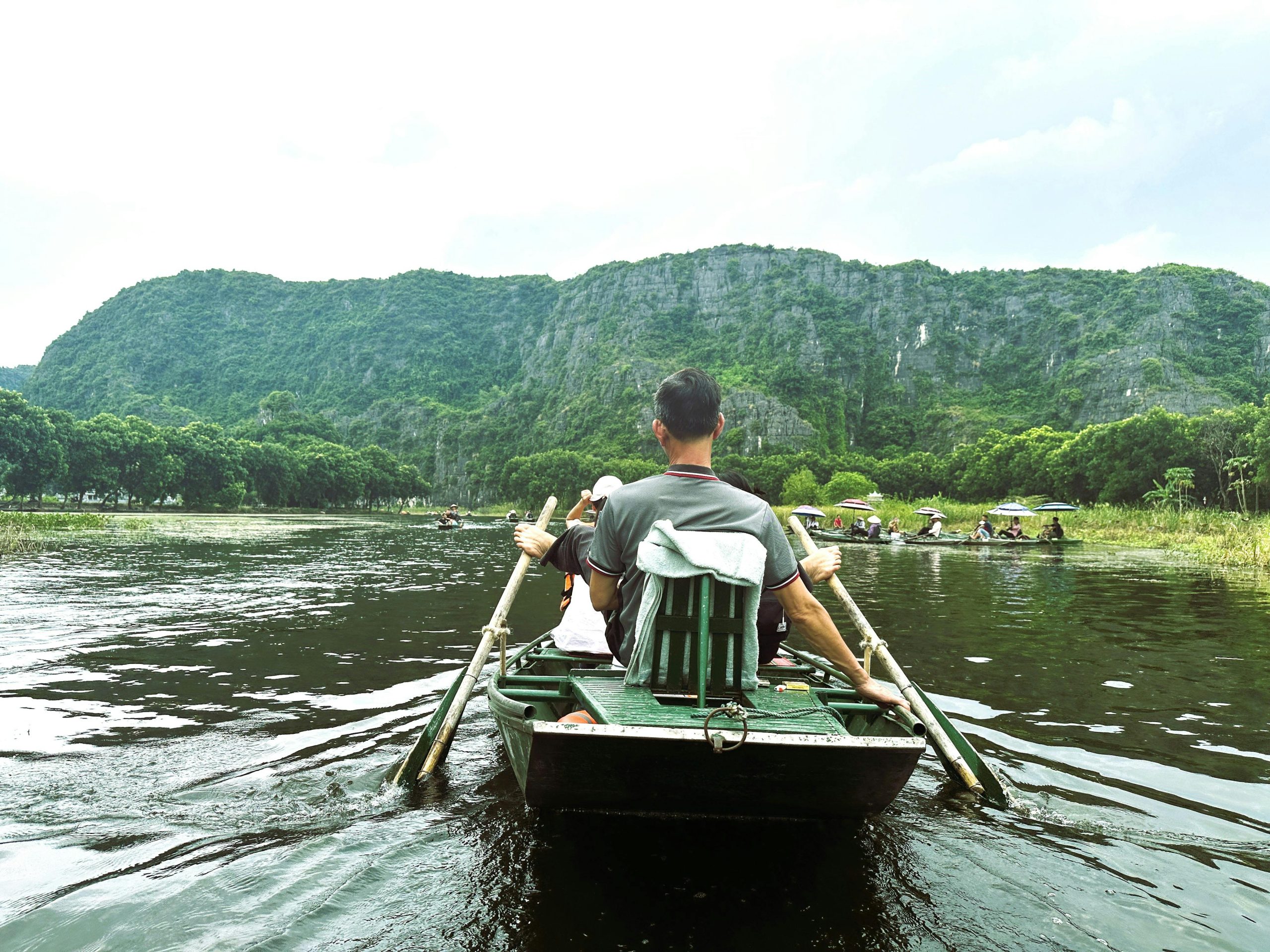 A traditional Vietnamese sampan boat being rowed by a woman using her feet, gliding through emerald rice paddies with towering limestone karsts in the background, on the Ngo Dong River in Tam Coc.