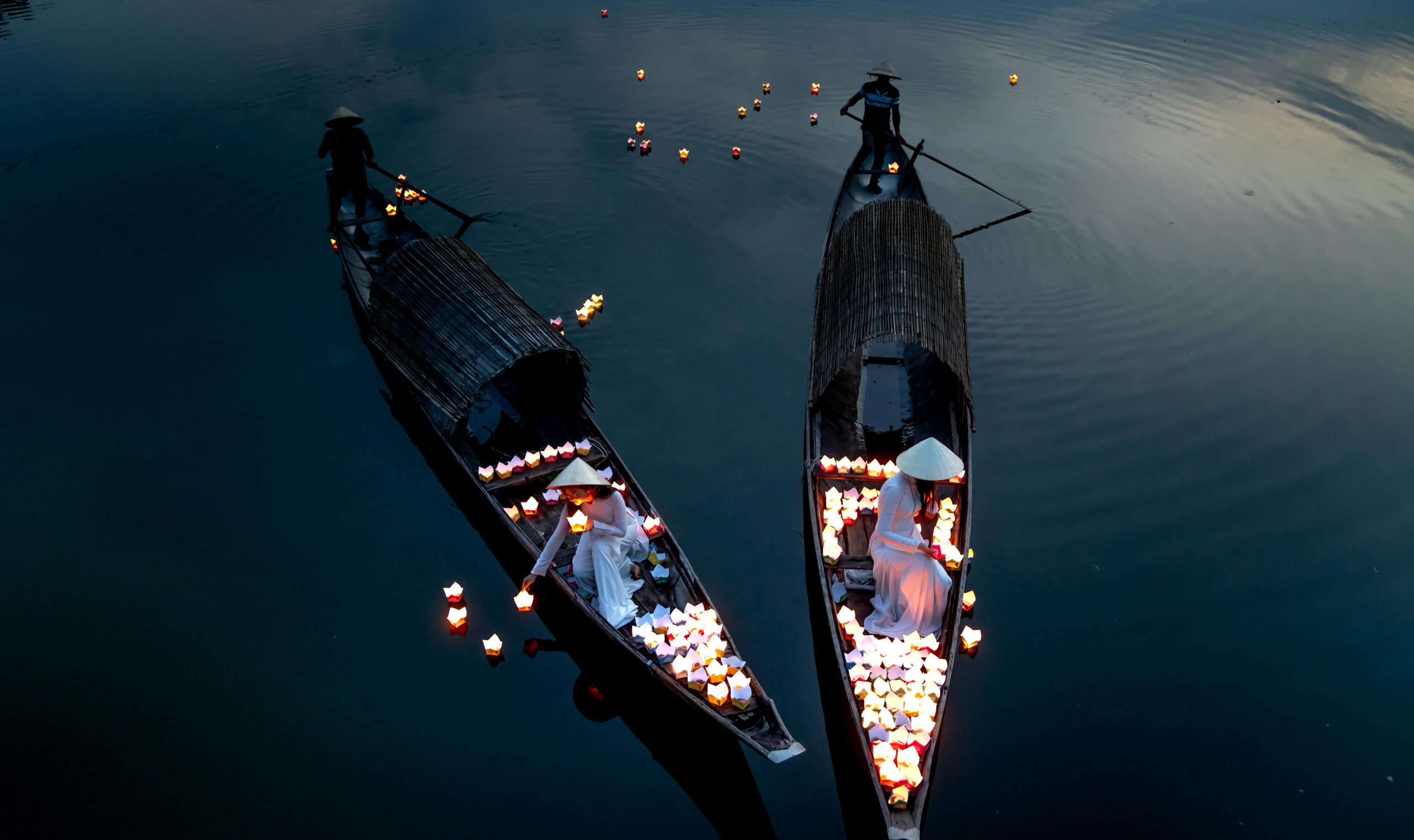 A traditional wooden boat adorned with lit colorful lanterns gently floating down the Thu Bon River in Hoi An at twilight. The river reflects the warm glow of the lanterns and the soft orange sky.