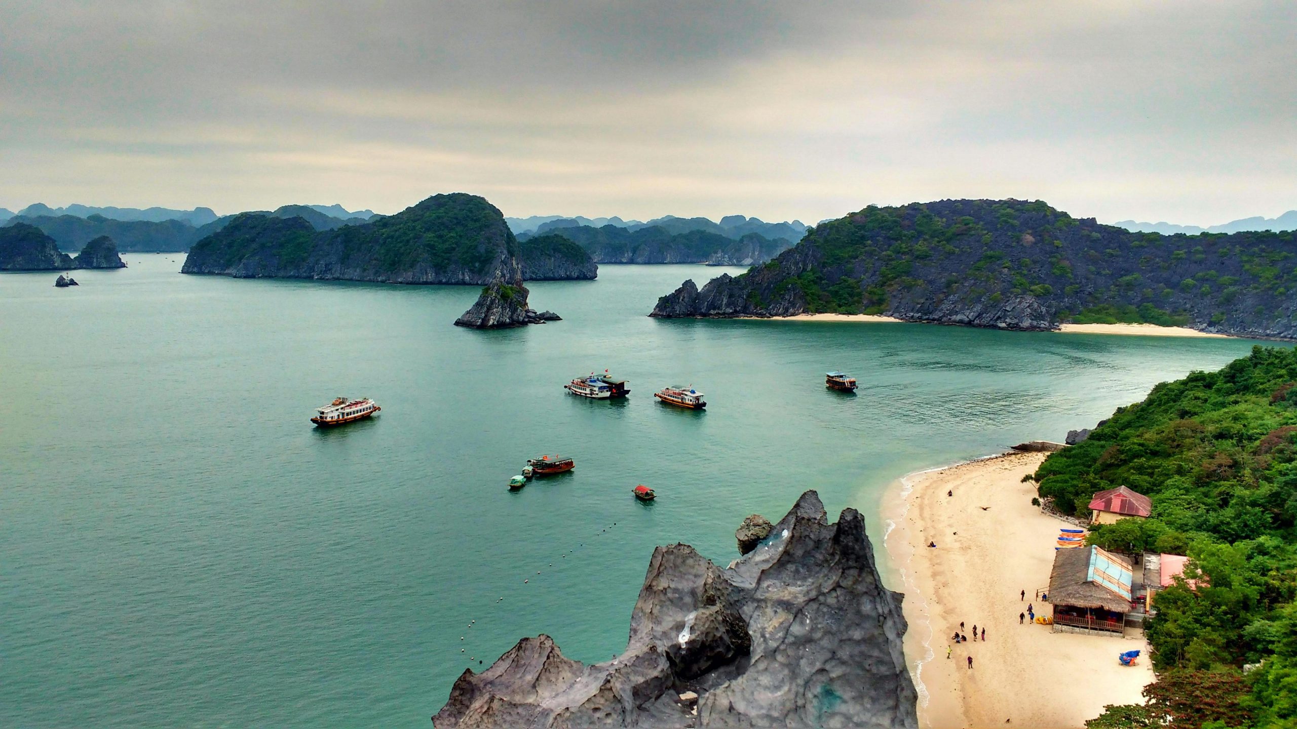A traditional wooden junk boat with red sails gracefully navigating through the calm emerald waters of Ha Long Bay, surrounded by numerous towering, lush green limestone karsts under a bright blue sky