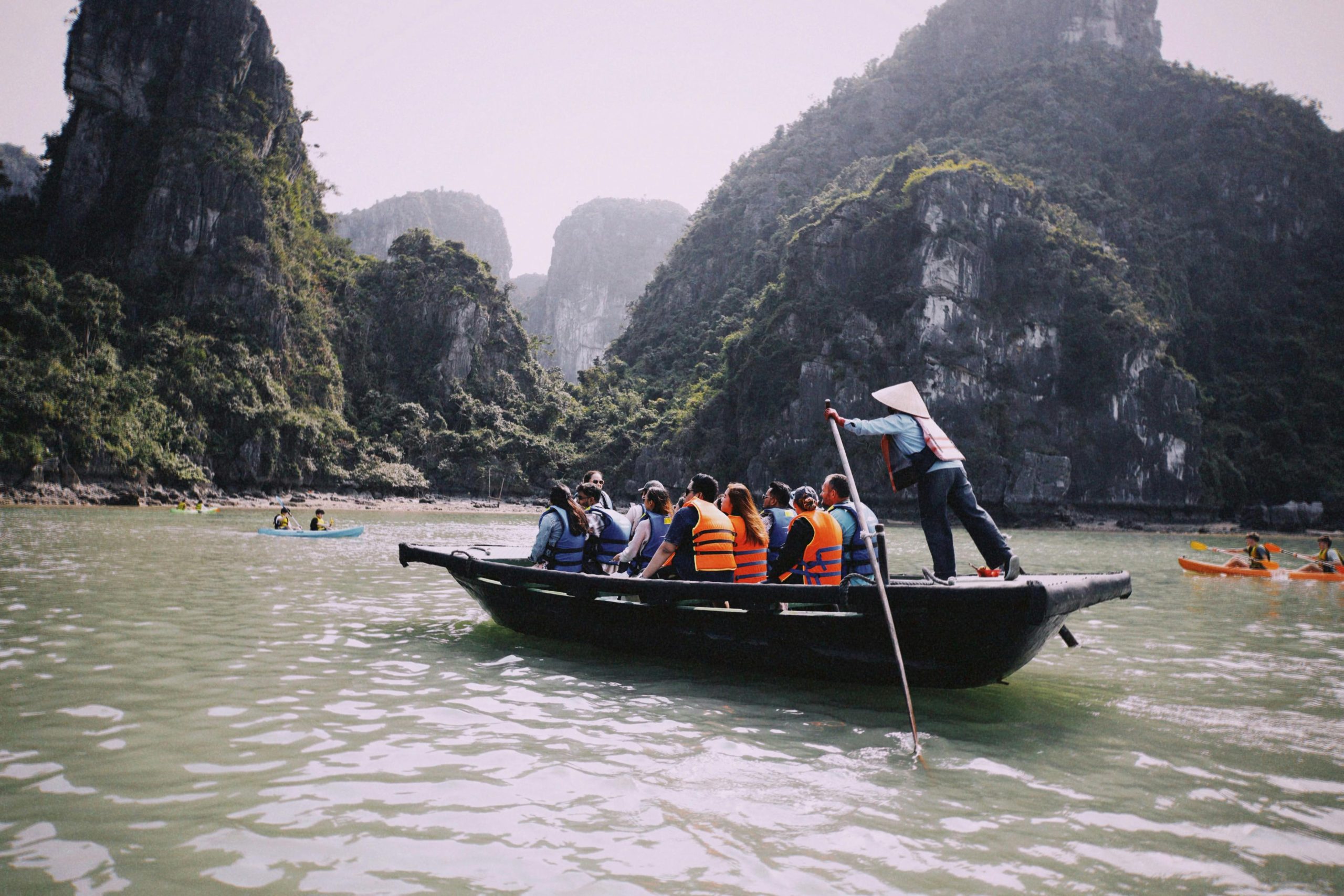 A traditional wooden sampan boat with a local rower navigating through calm emerald waters, flanked by towering limestone karsts covered in lush green foliage under a clear blue sky in Trang An Grottoes, Ninh Binh.
