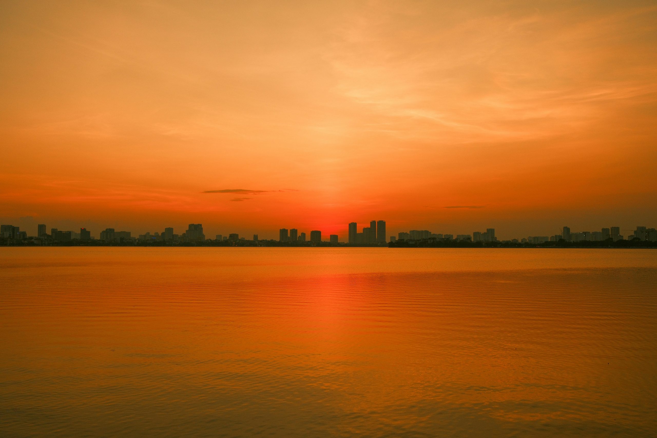 A tranquil sunrise over Hanoi's West Lake (Tay Ho), featuring calm waters reflecting the early morning light and distant city skyline with hints of mist.