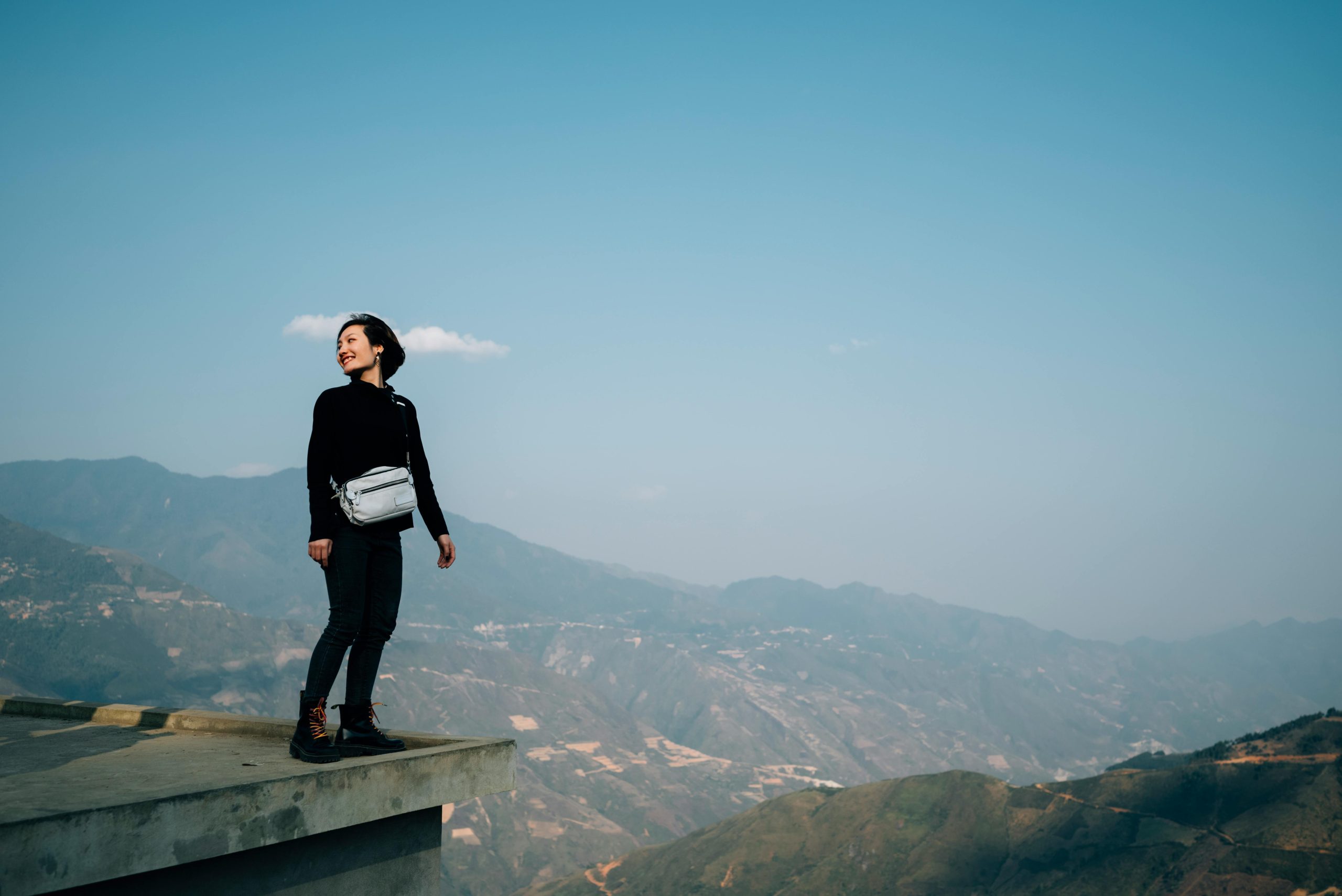 A traveler wearing warm clothing and a backpack stands at the summit of Fansipan Peak, looking out at a panoramic view of layered mountain ranges disappearing into the distance under a clear sky