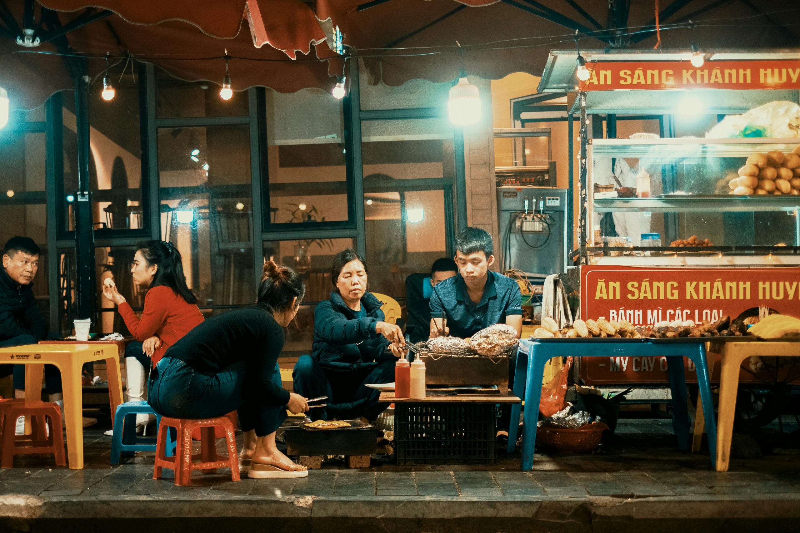 A vibrant, close-up shot of various colorful Vietnamese street food dishes laid out on a stall in Ben Thanh Market at night, with blurred people in the background enjoying the ambiance