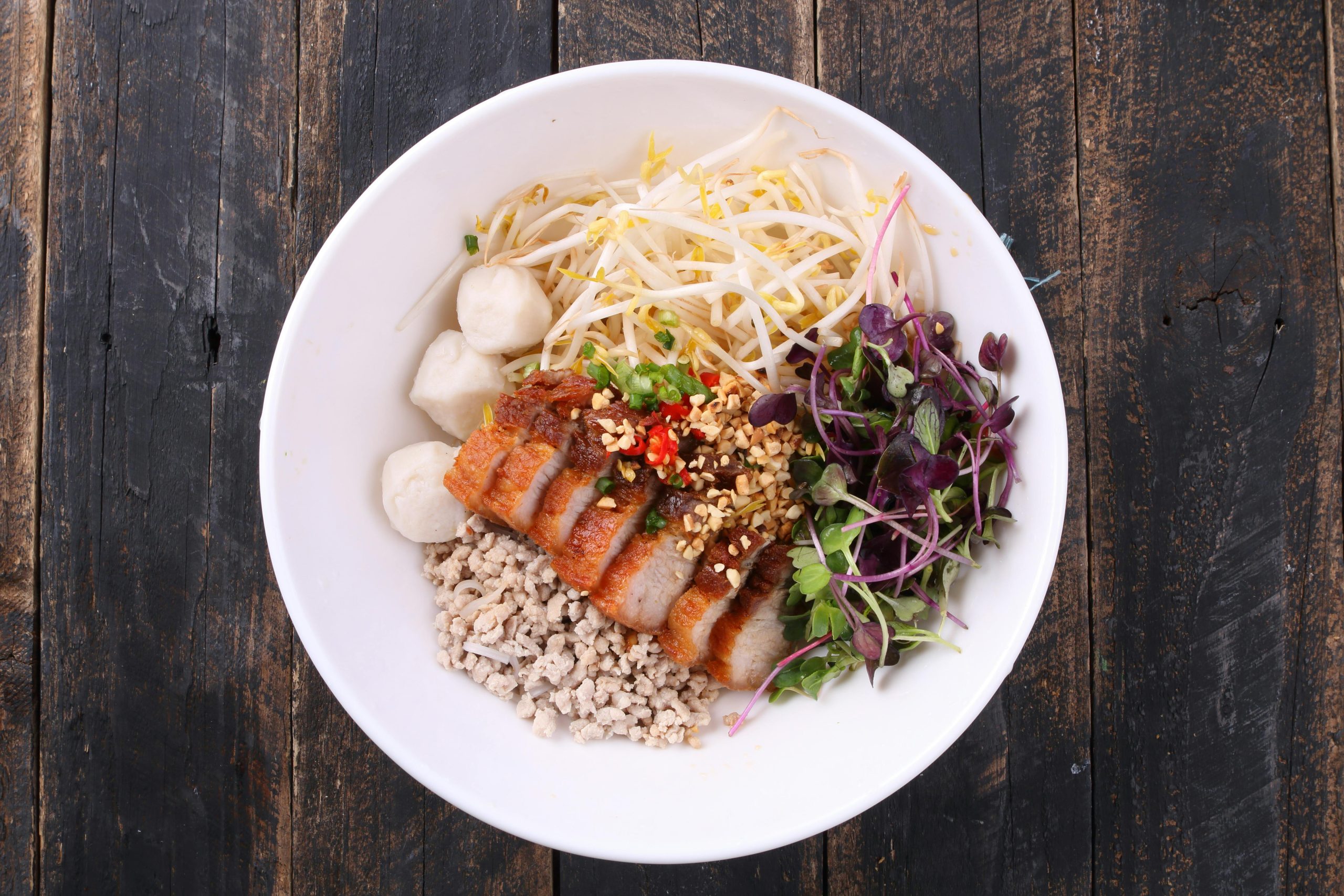 A vibrant Vietnamese dish called Cao Lau, featuring thick noodles, slices of roasted pork, fresh green herbs, and crispy rice crackers, served in a traditional bowl on a rustic wooden table in a Hoi An restaurant.