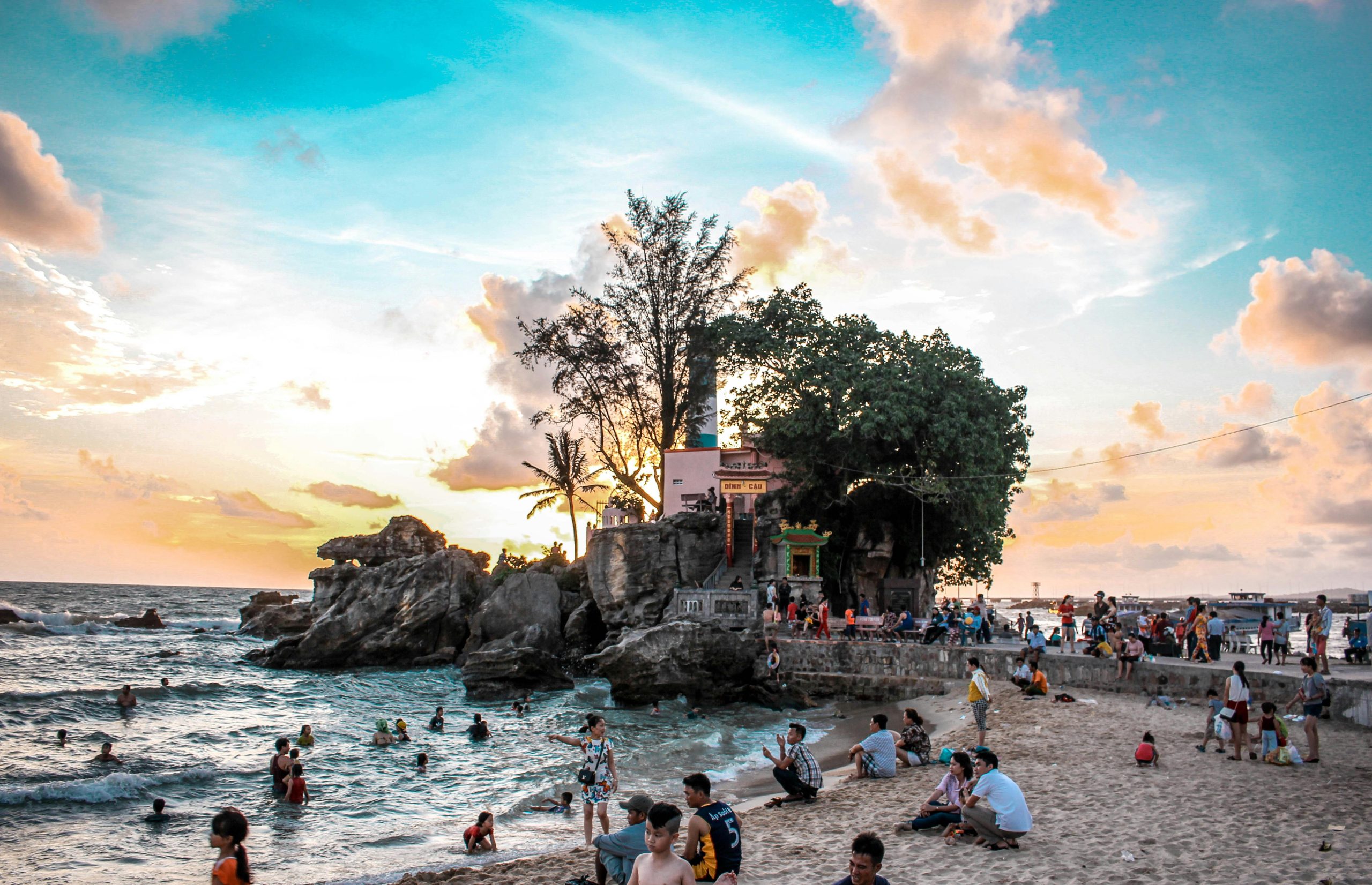A wide shot of Long Beach (Bai Truong) in Phu Quoc, Vietnam, at sunset, with golden sand, calm turquoise water, silhouetted palm trees, and a few people enjoying the view.