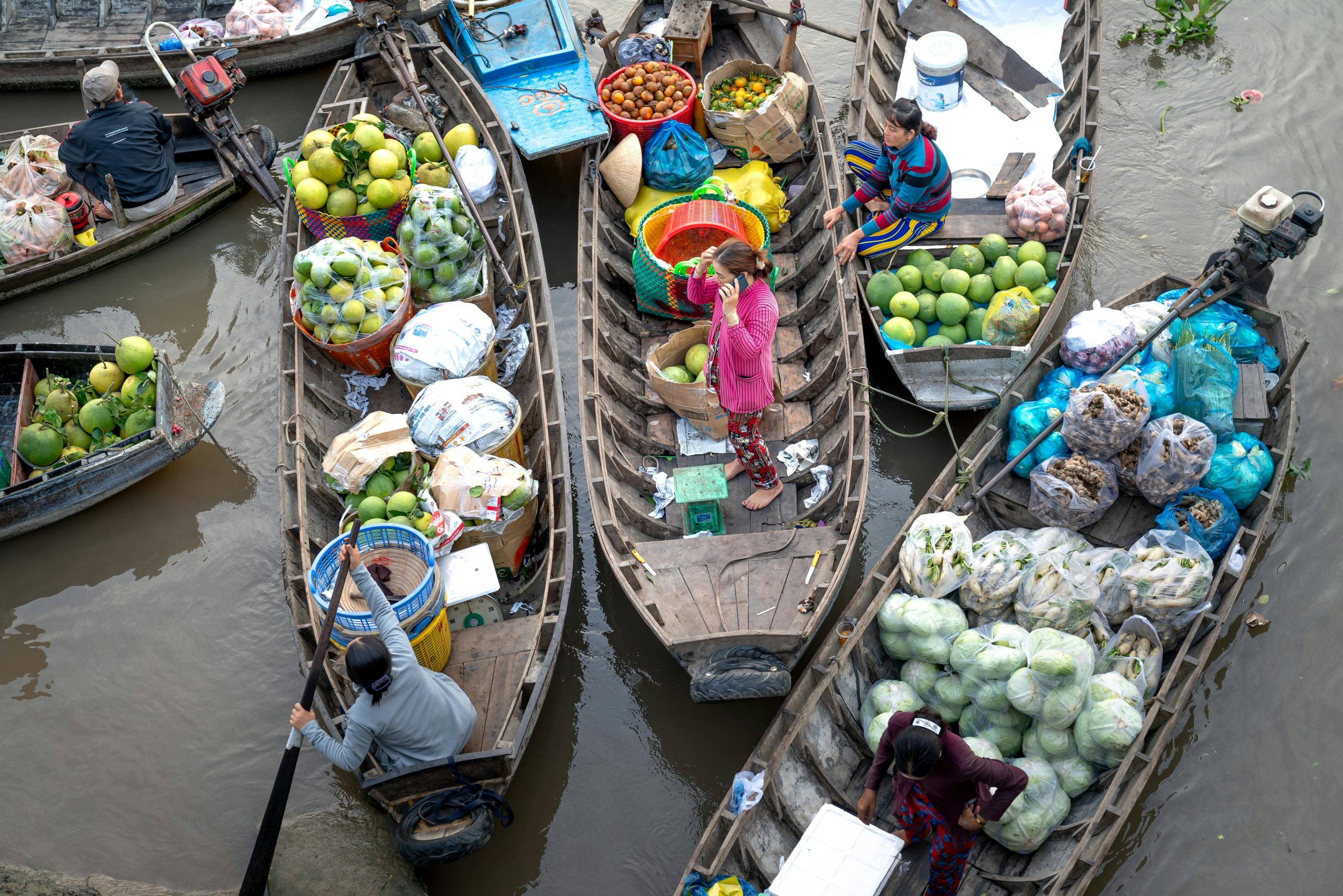 A wide shot of the Cai Rang Floating Market at sunrise, with many sampan boats and larger vessels selling a variety of colorful fruits and vegetables, people interacting, and a calm river reflecting the morning sky.