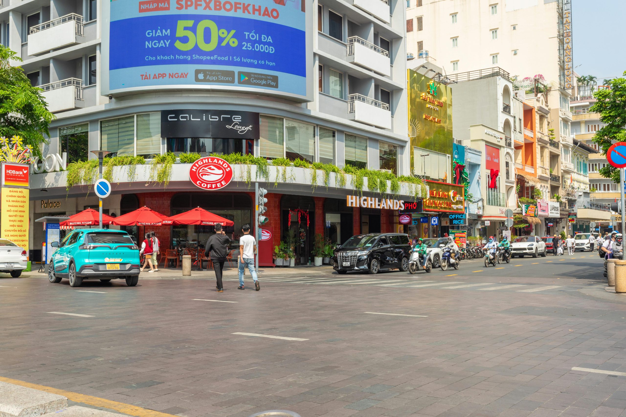 A wide shot of the Notre Dame Cathedral Basilica of Saigon and the Central Post Office in District 1, Ho Chi Minh City, under a clear blue sky with motorbikes and pedestrians in the foreground
