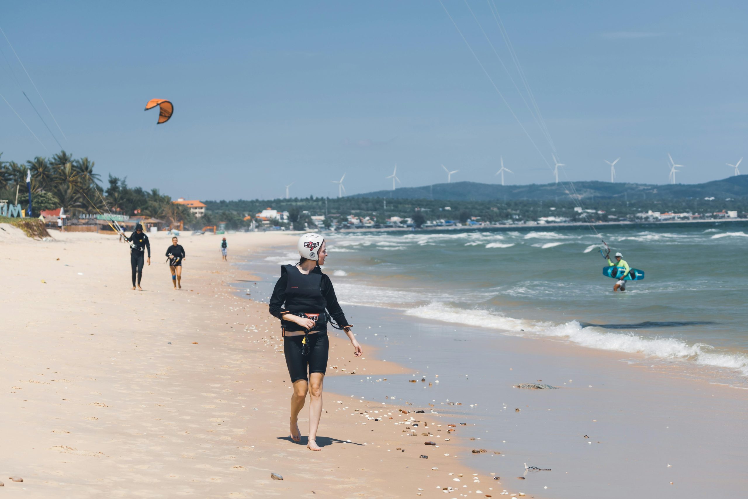 Multiple colorful kitesurfers in action on the turquoise waters of Mui Ne Beach, with a backdrop of a sandy beach and palm trees under a bright blue sky.