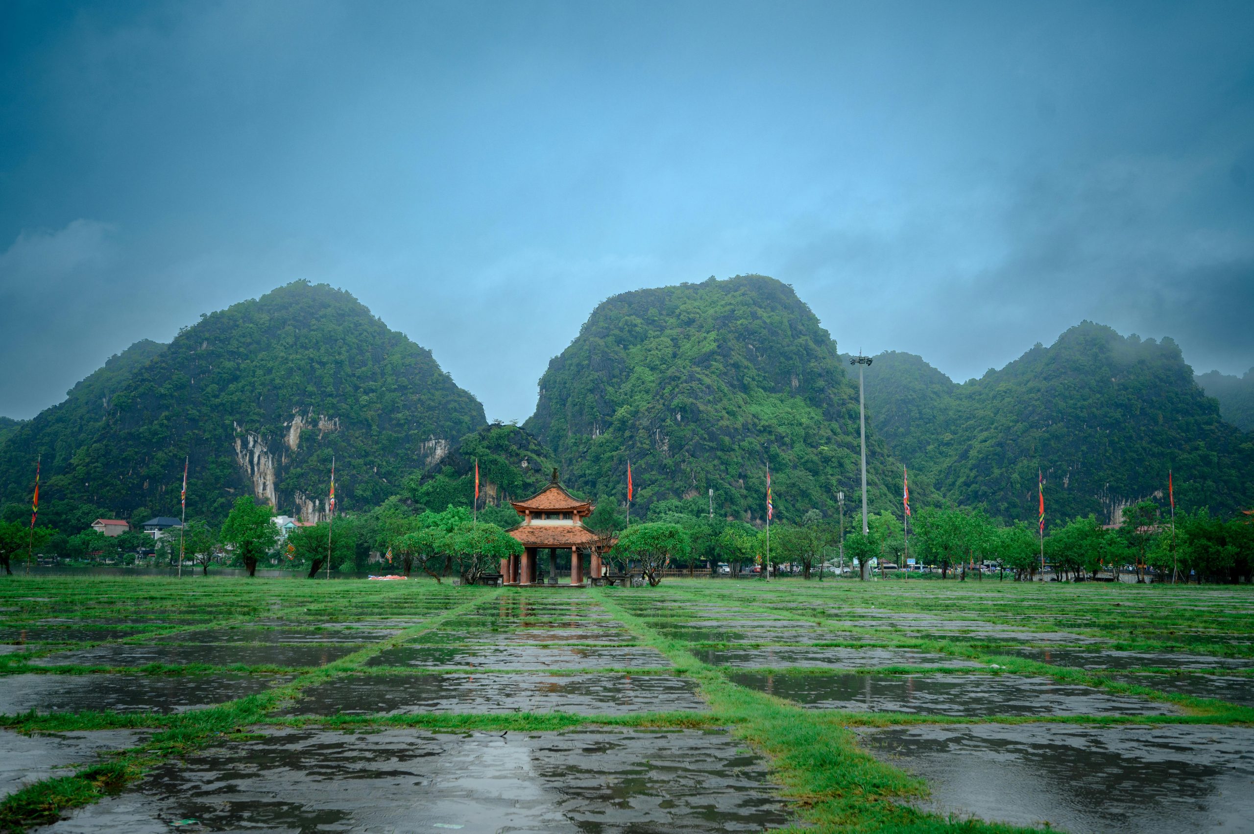 Panoramic view of Hoa Lu Ancient Capital surrounded by lush green rice fields and towering limestone mountains under a bright blue sky, showcasing the natural defenses and scenic beauty