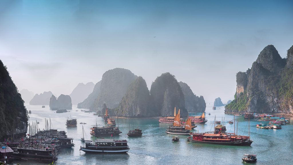 A luxurious traditional wooden junk boat with red sails cruising through the calm, emerald waters of Ha Long Bay, surrounded by towering limestone karsts under a clear blue sky.