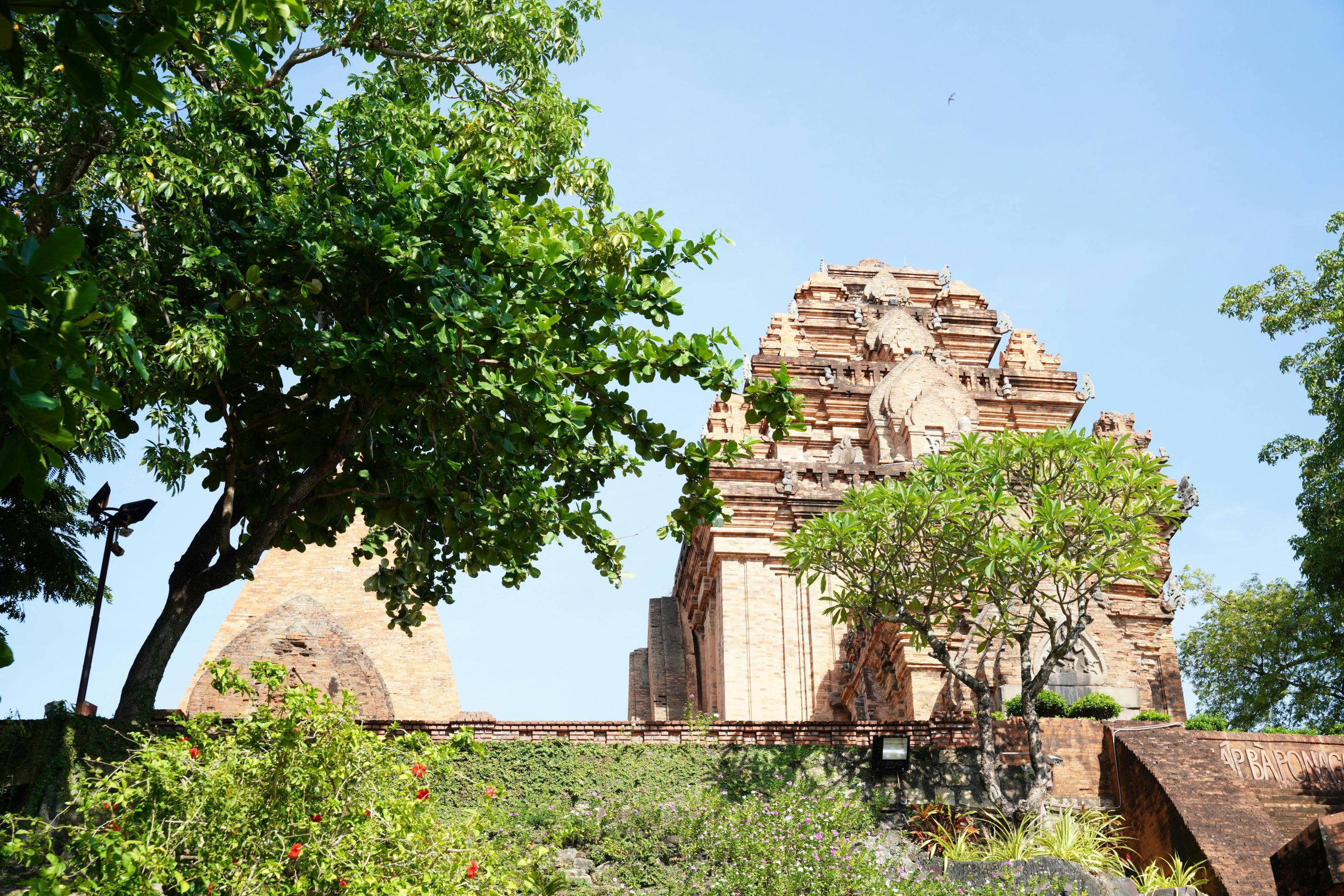 The ancient brick towers of Po Nagar Cham complex on a sunny day, with intricate carvings visible and green trees surrounding the historical site.