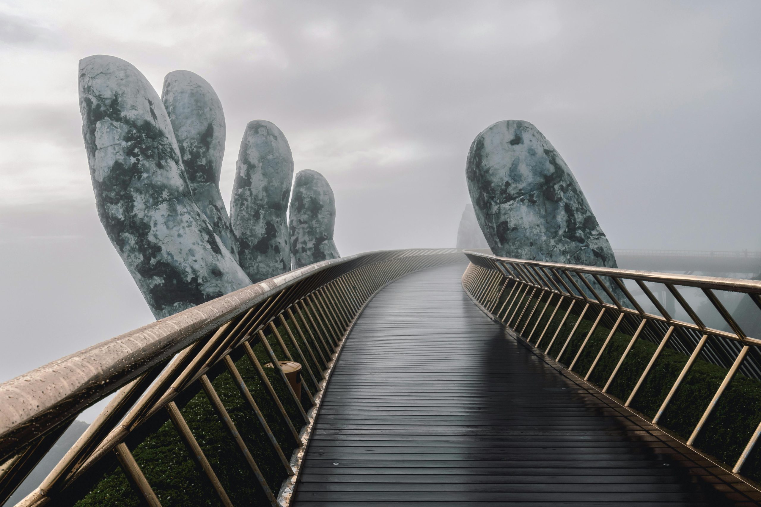 The iconic Golden Bridge at Ba Na Hills, supported by two colossal weathered stone hands, with people walking on it and lush green mountains shrouded in mist in the background