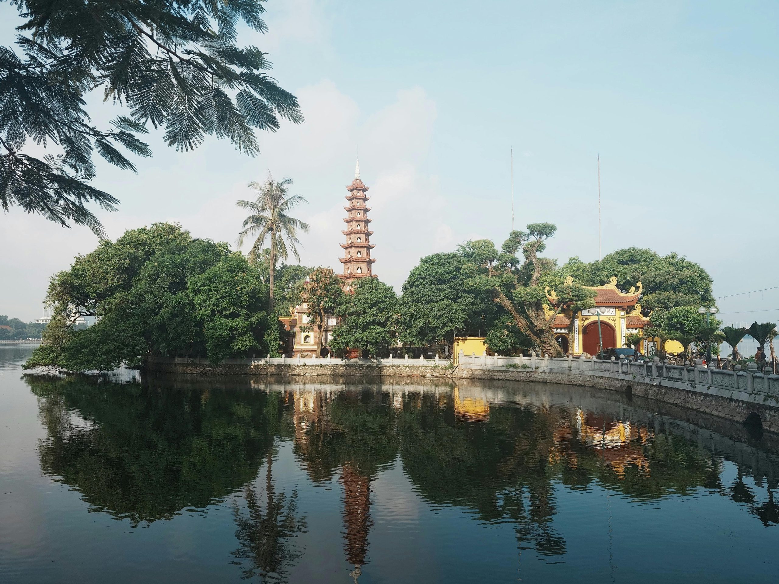 The iconic multi-tiered stupa of Tran Quoc Pagoda, beautifully situated on an islet in West Lake, surrounded by lush greenery and calm water, under a clear blue sky.