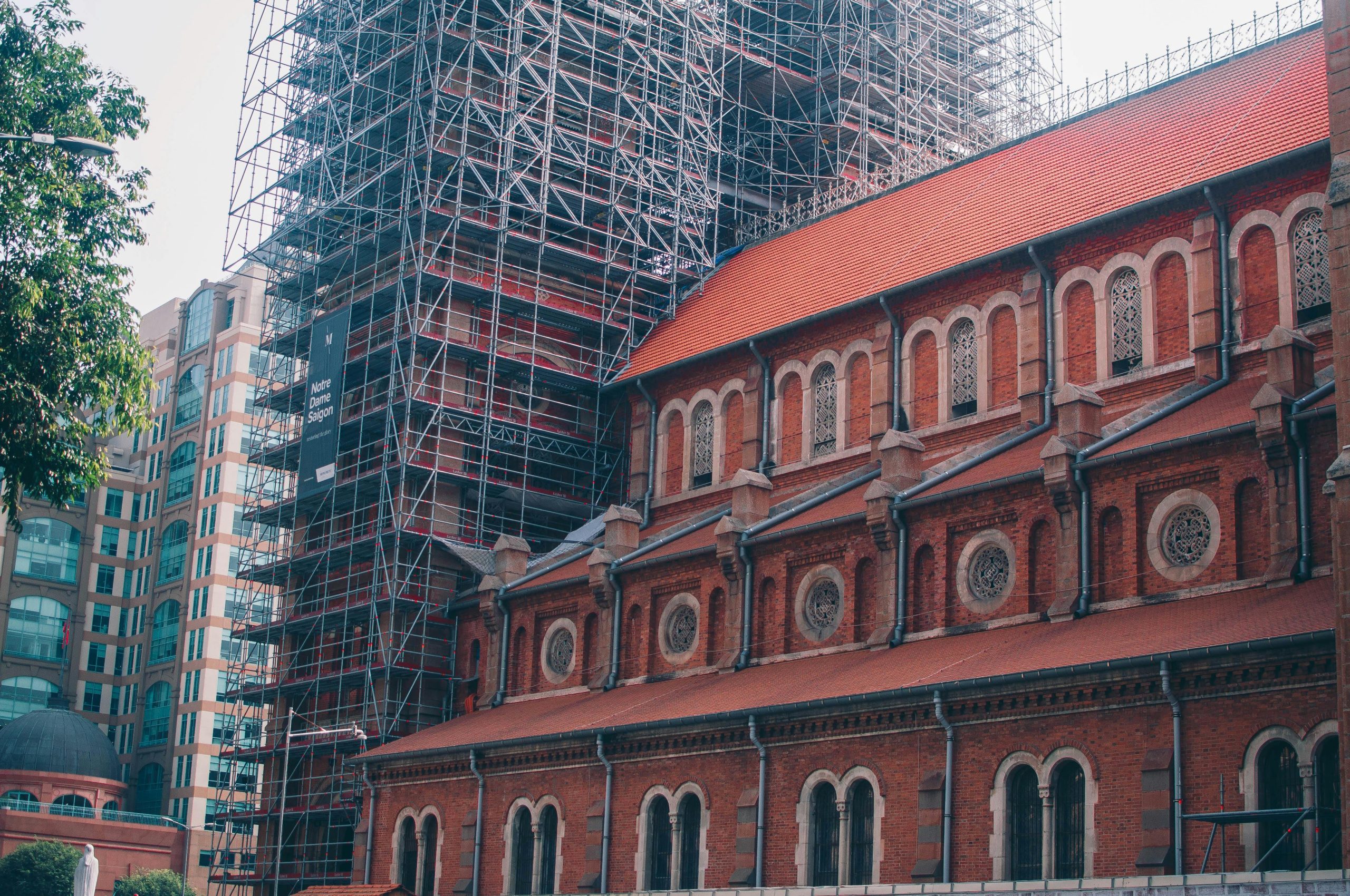 The iconic Notre Dame Cathedral Basilica of Saigon with its distinctive red brick facade under a bright blue sky, surrounded by green trees and bustling city life in Ho Chi Minh City, Vietnam.
