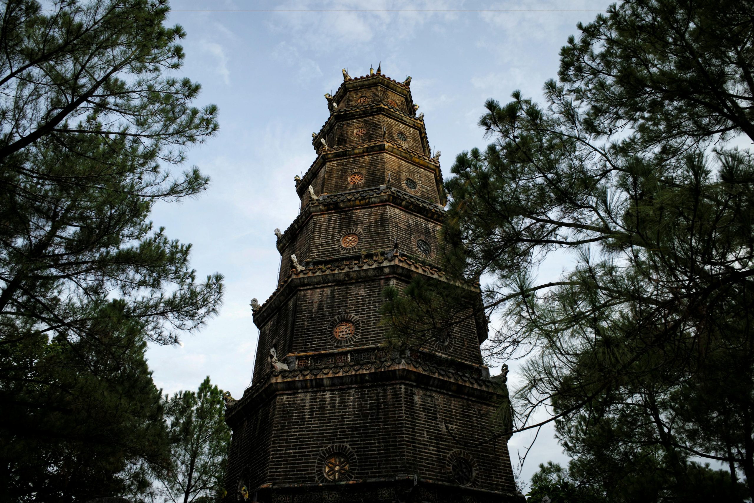 The iconic seven-story Phuoc Duyen Tower of Thien Mu Pagoda, a traditional Vietnamese pagoda, standing tall against a bright blue sky. The Perfume River is visible in the foreground with a traditional boat, and lush green trees surround the pagoda.