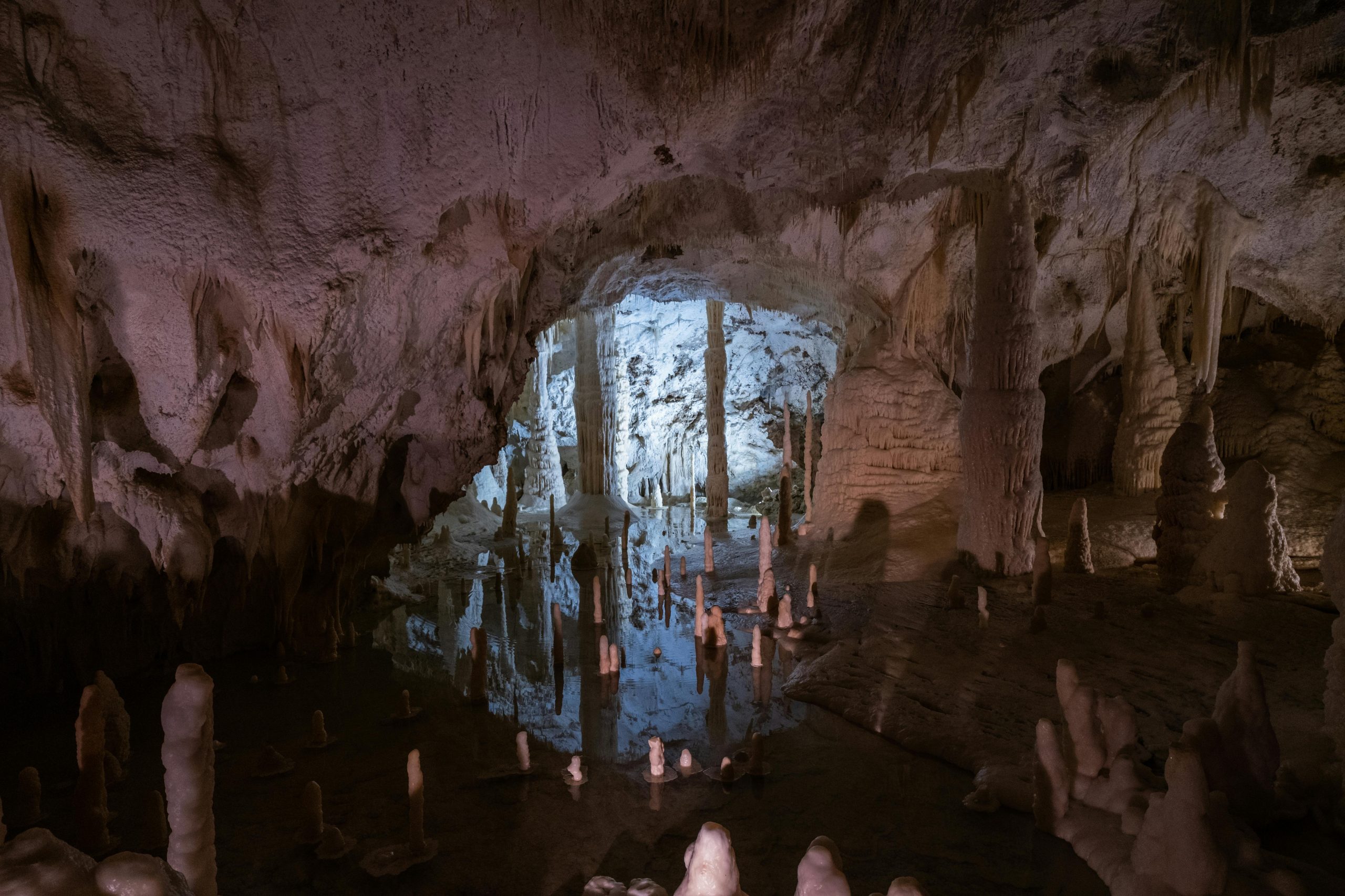 The interior of a dark, majestic cave in Trang An Grottoes, with faint light filtering in from the entrance and reflecting off the smooth water surface, showing ancient stalactite formations.