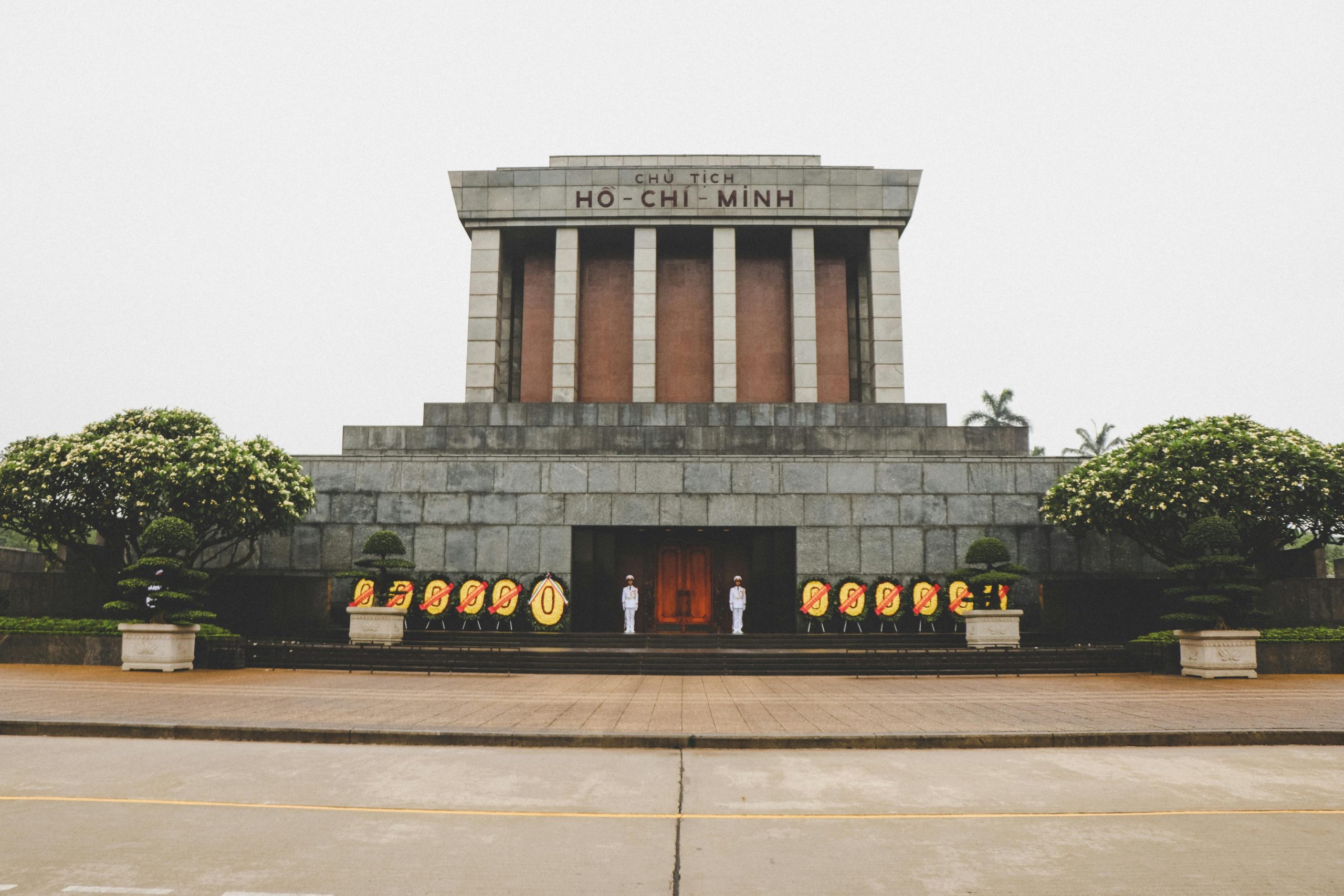 The majestic Ho Chi Minh Mausoleum at dawn, with the large Ba Dinh Square in front, illuminated by soft morning light, and a few people walking respectfully