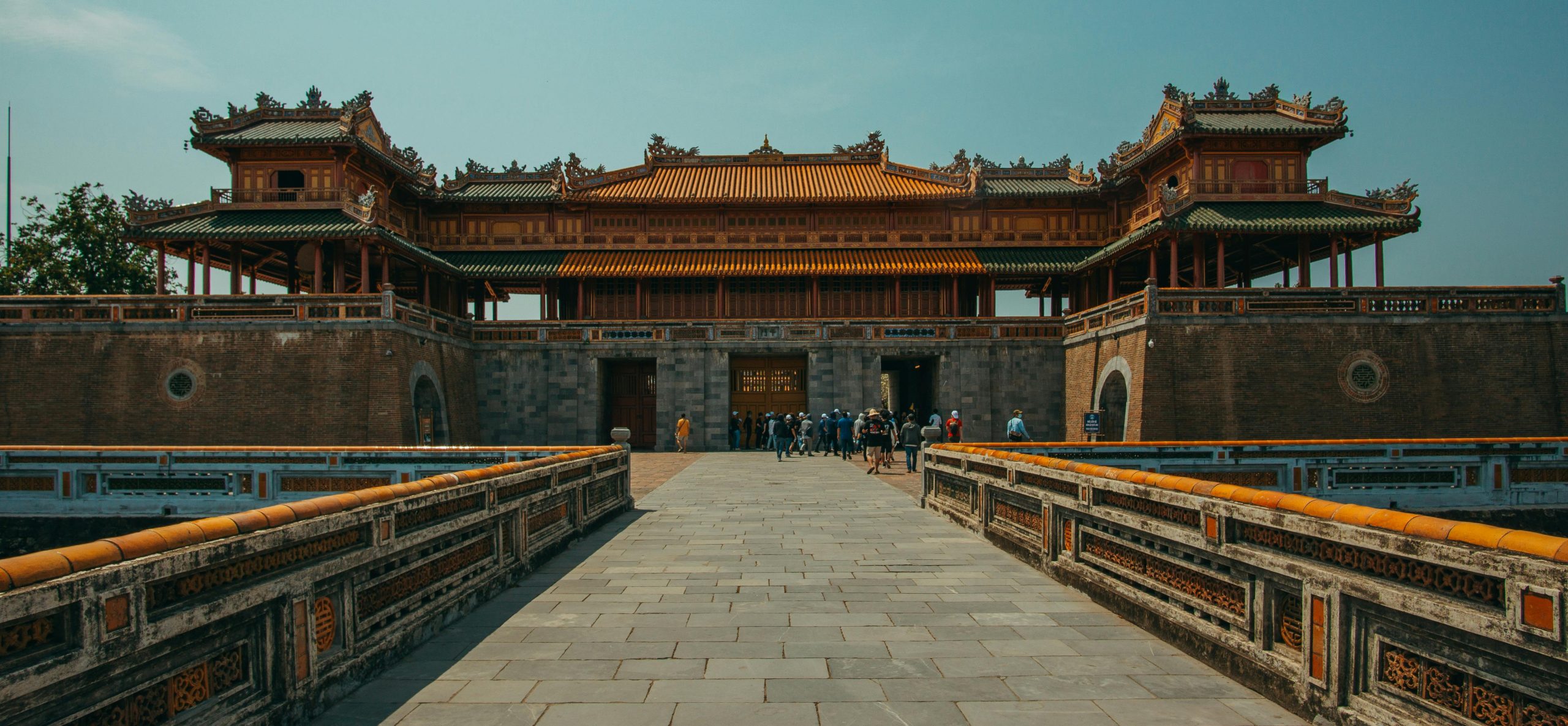 The Ngo Mon Gate (Meridian Gate) of the Hue Imperial City, showing intricate traditional Vietnamese architecture with a clear blue sky and lush surrounding gardens.