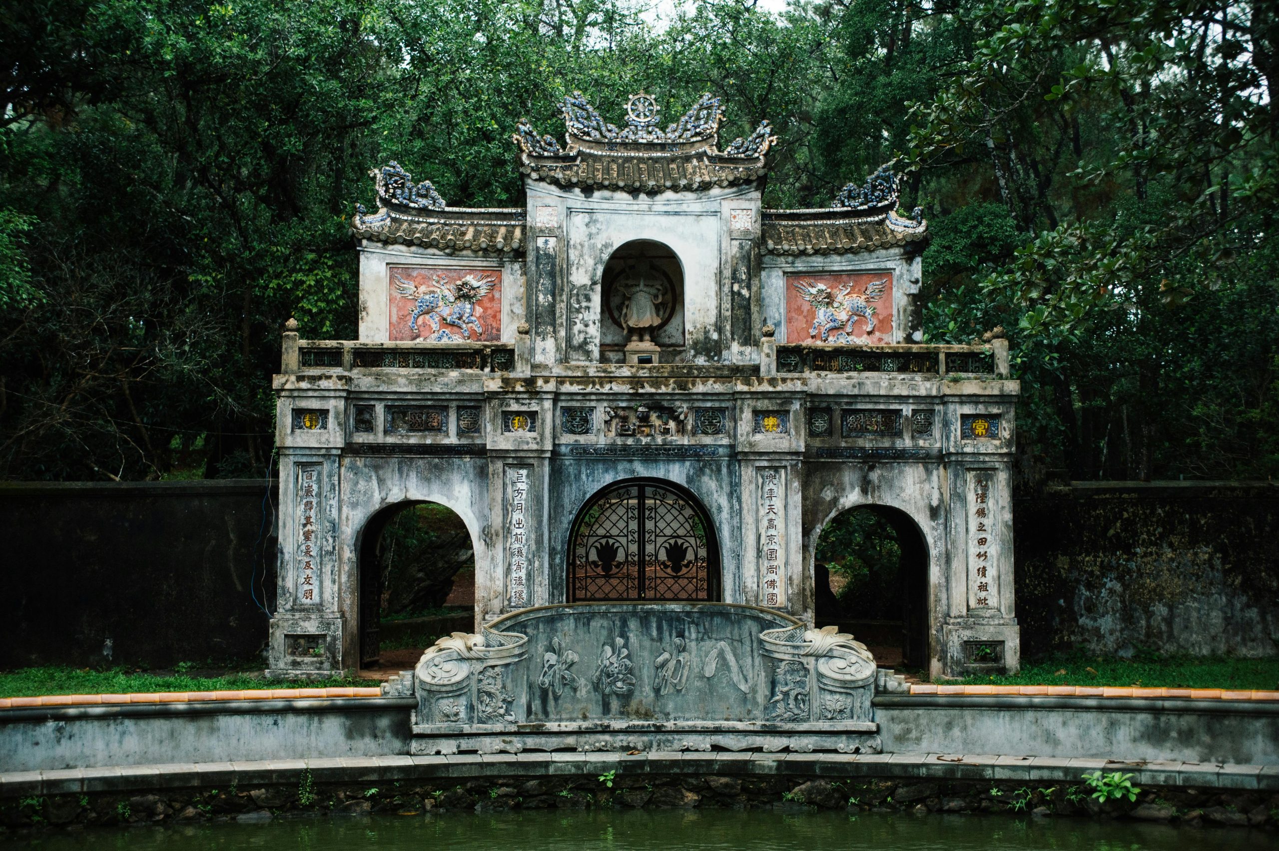 The ornate entrance gate of Bich Dong Pagoda, an ancient temple complex built into a cave, surrounded by lush green trees and reflecting in a calm lotus pond, with traditional Vietnamese architecture visible.