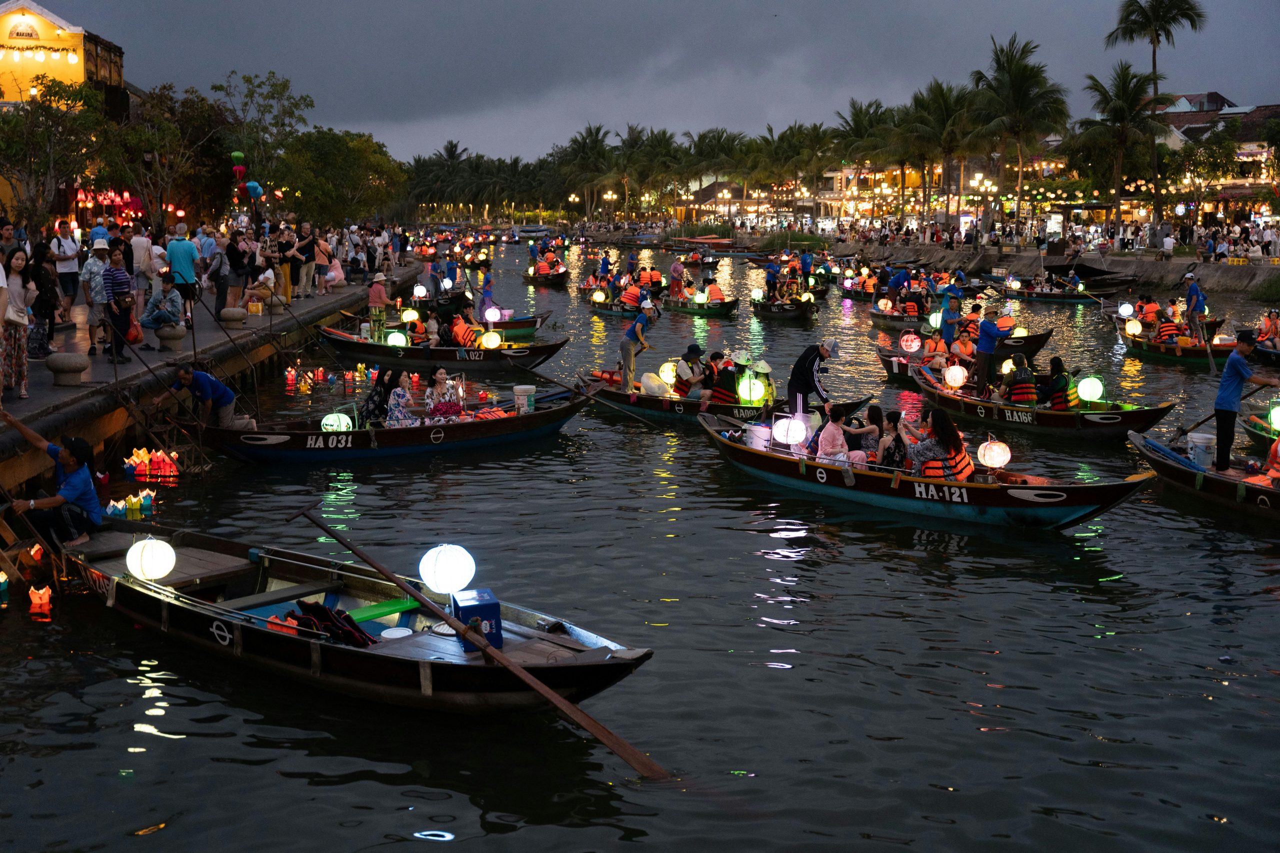 The vibrant and illuminated streets of Hoi An Ancient Town at dusk, with colorful lanterns hanging overhead, people strolling, and traditional wooden buildings lining the canals